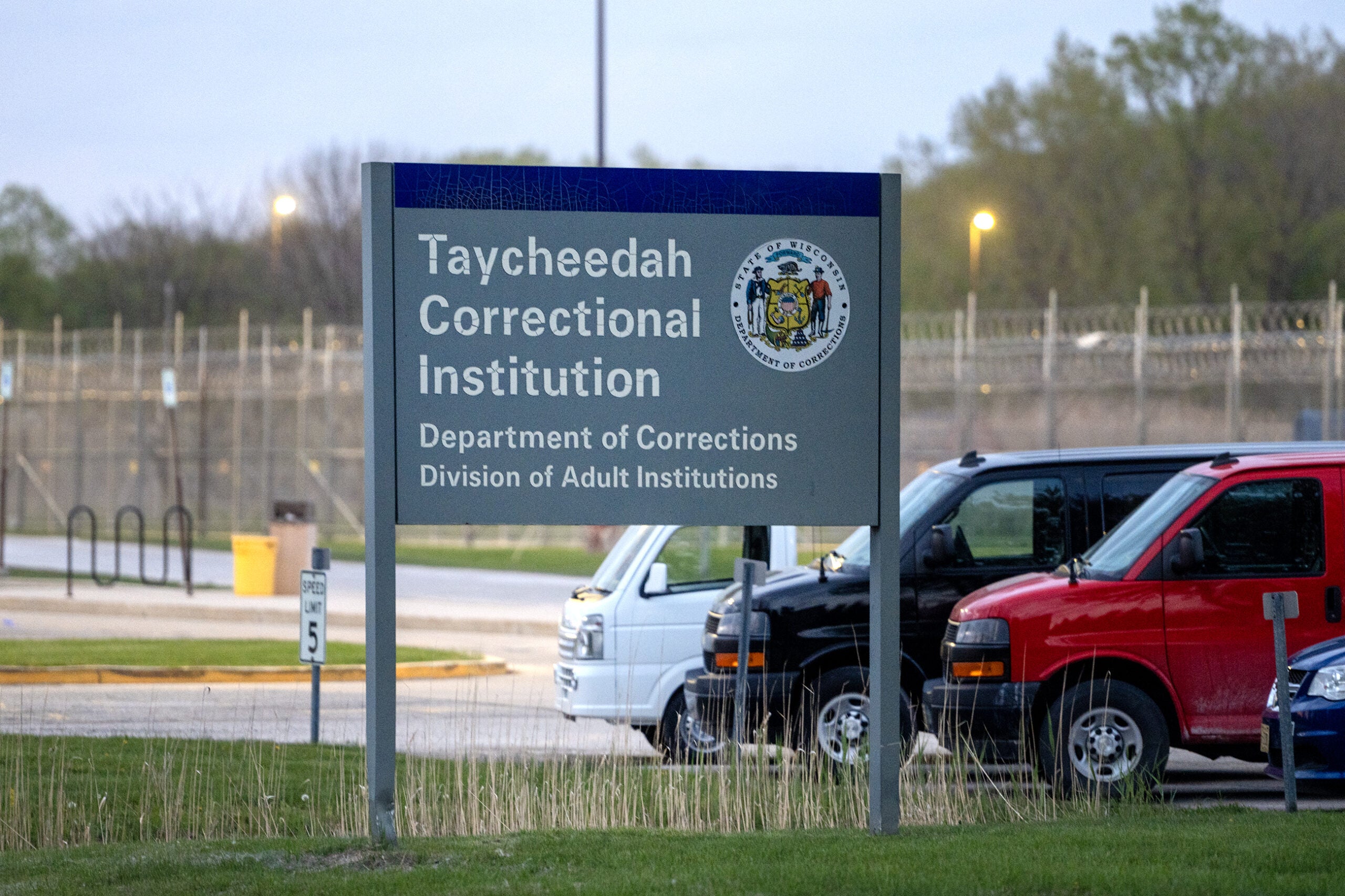 Sign for Taycheedah Correctional Institution stands in front of a parking lot with several vehicles and a fenced facility in the background.