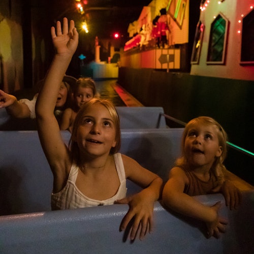 Children in a dark amusement ride; one girl raises her hand while others look excitedly at the animated scenes ahead.