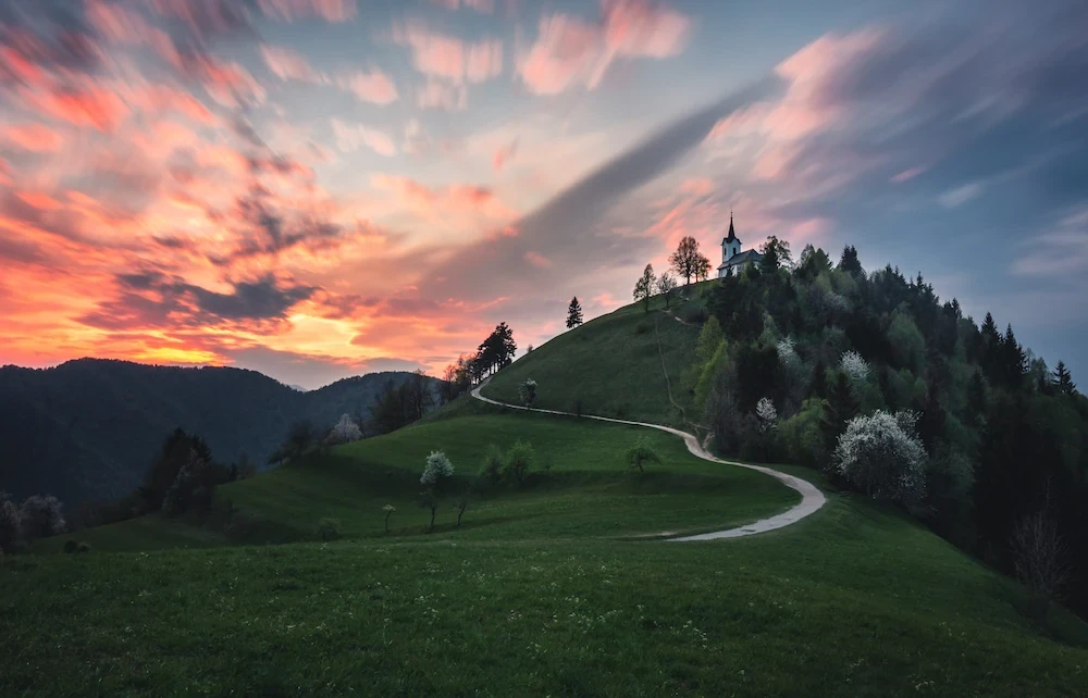 Sunset colored clouds above the hilltop church Sveti Jakob in Slovenia, with a winding path leading up the hill.