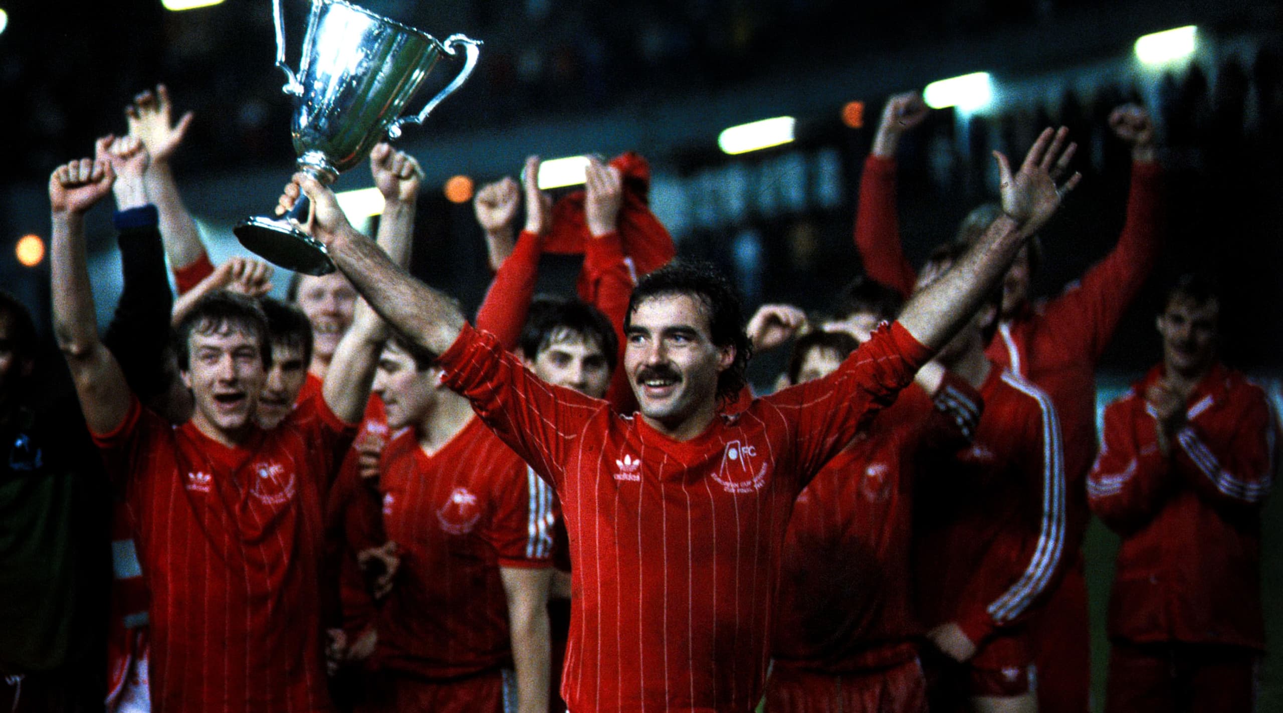 Man holding a cup in red football kit, with rest of team behind him