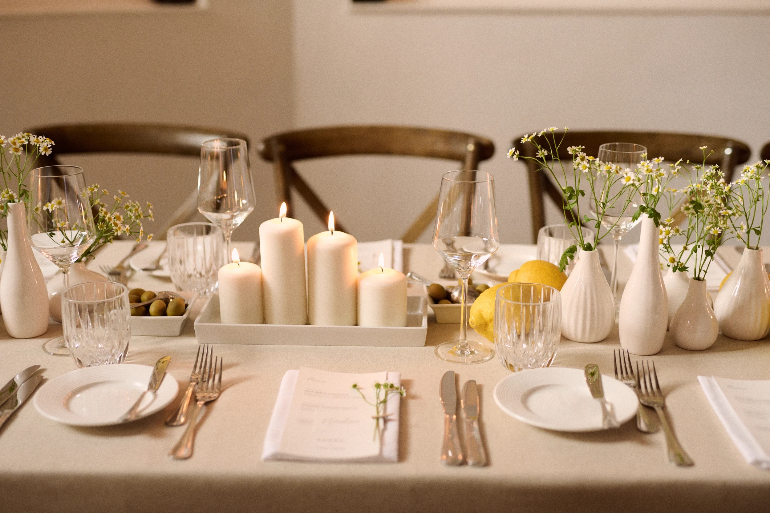 Table setting with pillar candles, daisies, olives and lemons on a linen tablecloth.