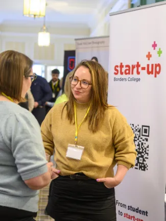 Two people interact at a networking event beside a "start-up Borders College" banner. The atmosphere is professional and engaging.