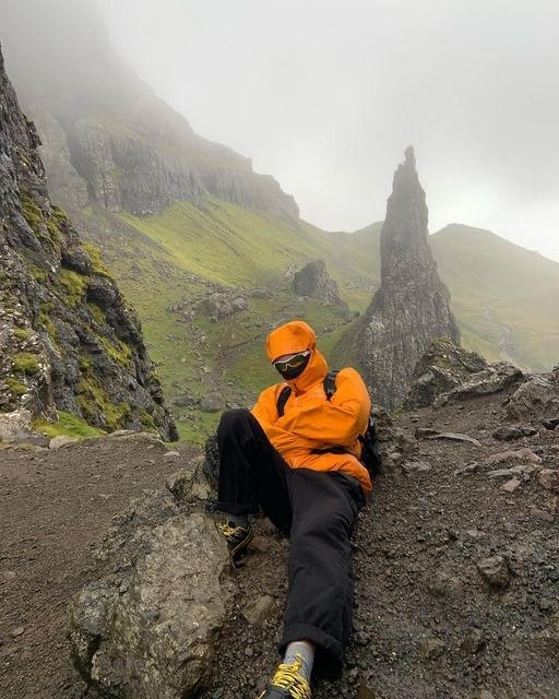A smiling woman with her arms crossed, standing against a dark green background. She has long, dark hair.