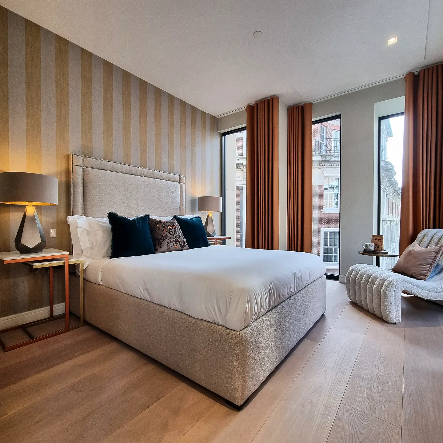 A modern, elegant bedroom features a plush king-sized bed with crisp white linens and navy accent pillows, set against a striped accent wall, with tall windows draped in rust-colored curtains, providing a view of the buildings outside.
