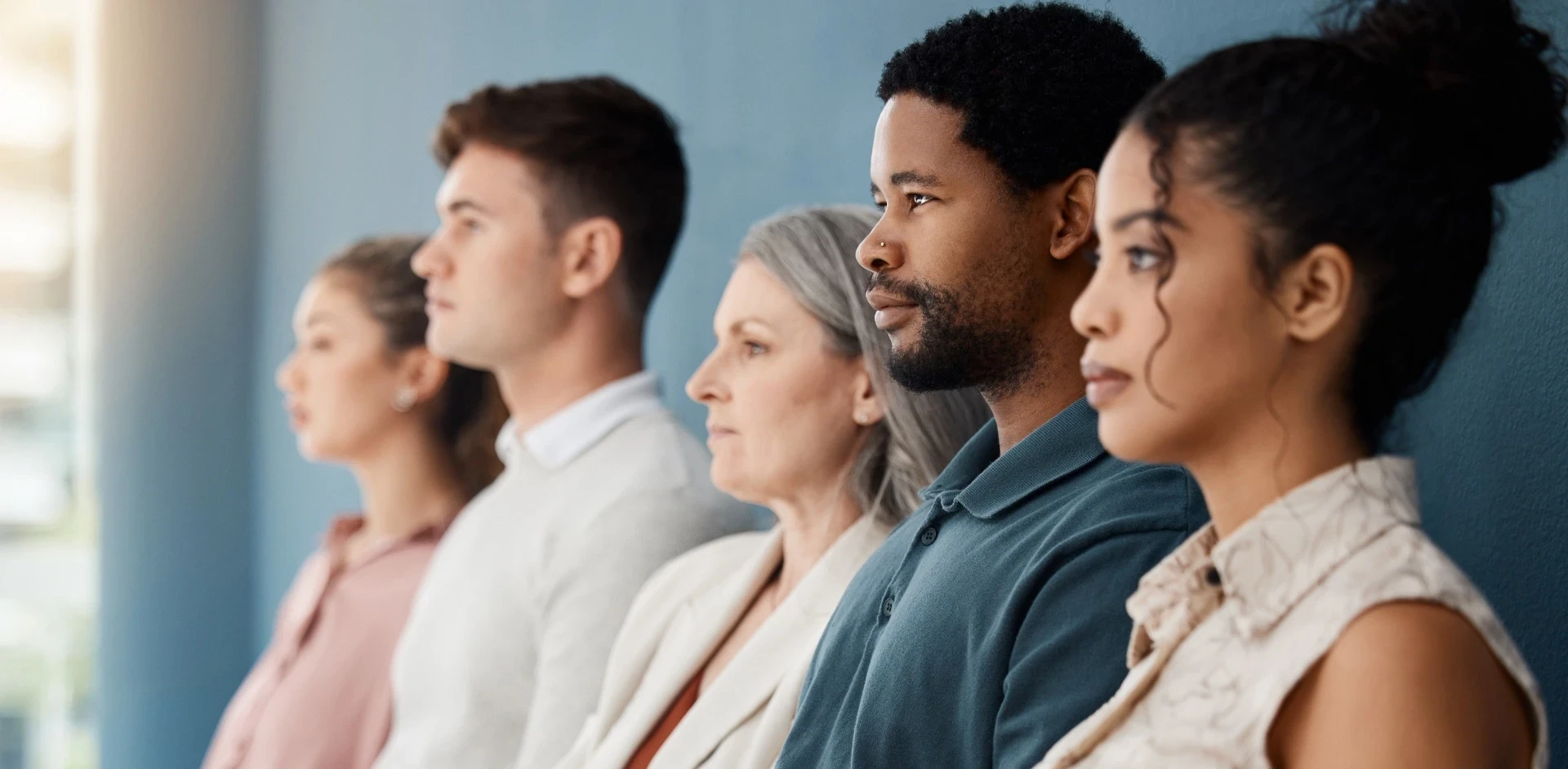 A imagem mostra um grupo diverso de pessoas em perfil, todas olhando na mesma direção, provavelmente para algo à frente. A diversidade é representada em idades, gêneros e etnias, ressaltando inclusão e igualdade. A luz suave do lado esquerdo da imagem ilumina os rostos, criando uma atmosfera serena e contemplativa. A composição transmite uma sensação de união e determinação compartilhada, como se todos estivessem comprometidos com um objetivo comum ou uma visão futura.