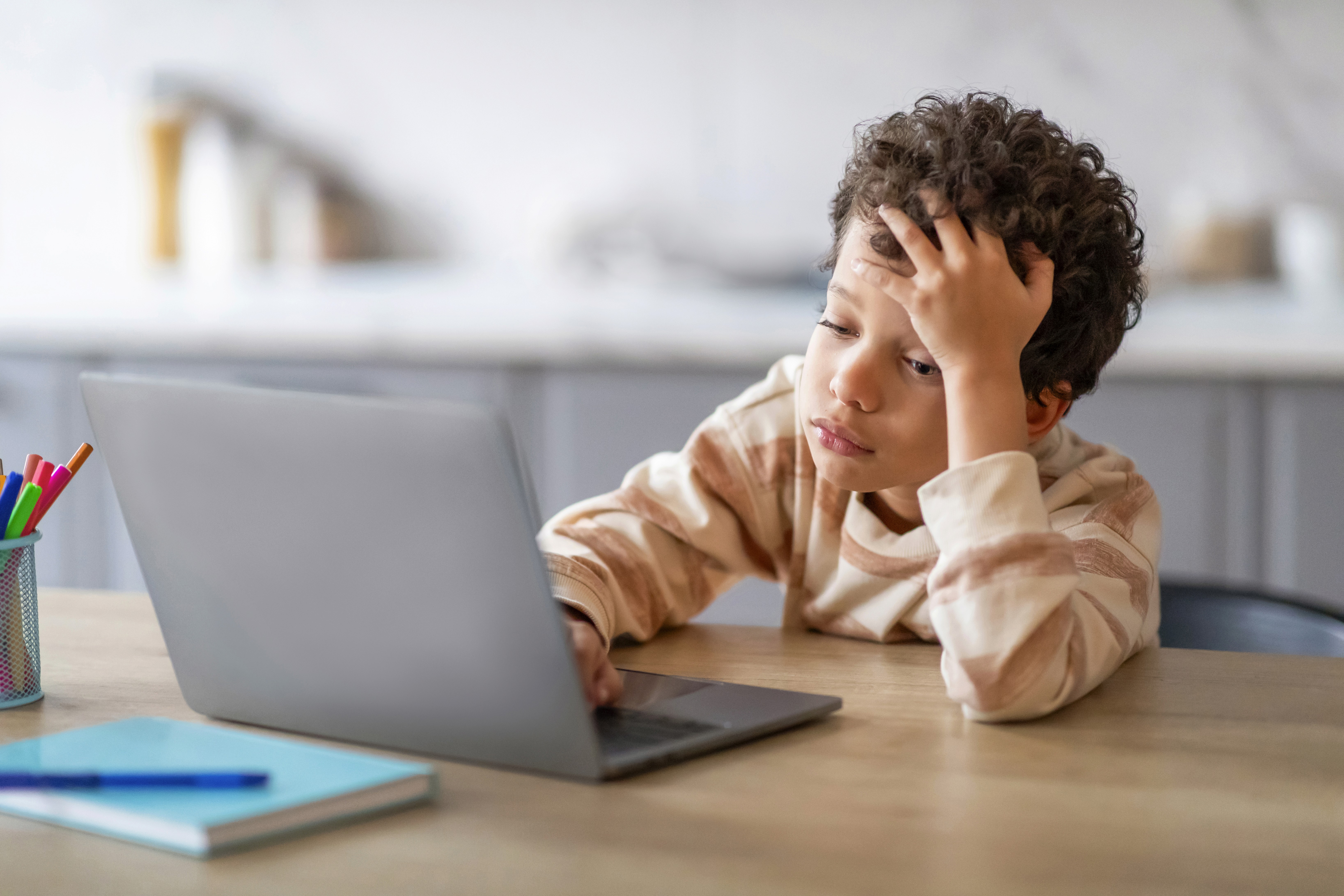 Criança sentada em frente ao notebook, apoiando a testa sobre uma das mãos, com expressão de tédio.