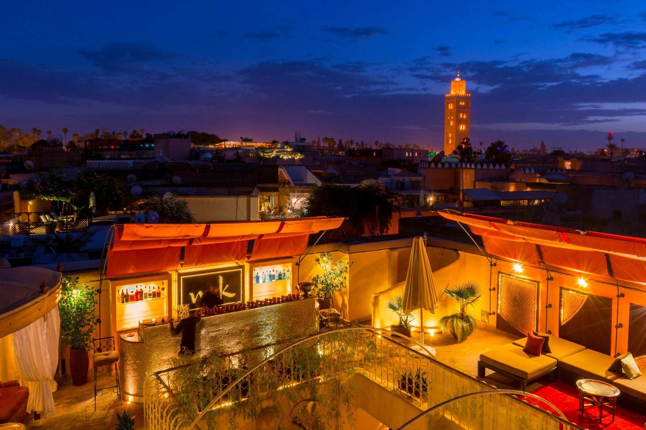 A rooftop terrace with orange and yellow lights overlooks a coastal town at dusk under a deep blue twilight sky.