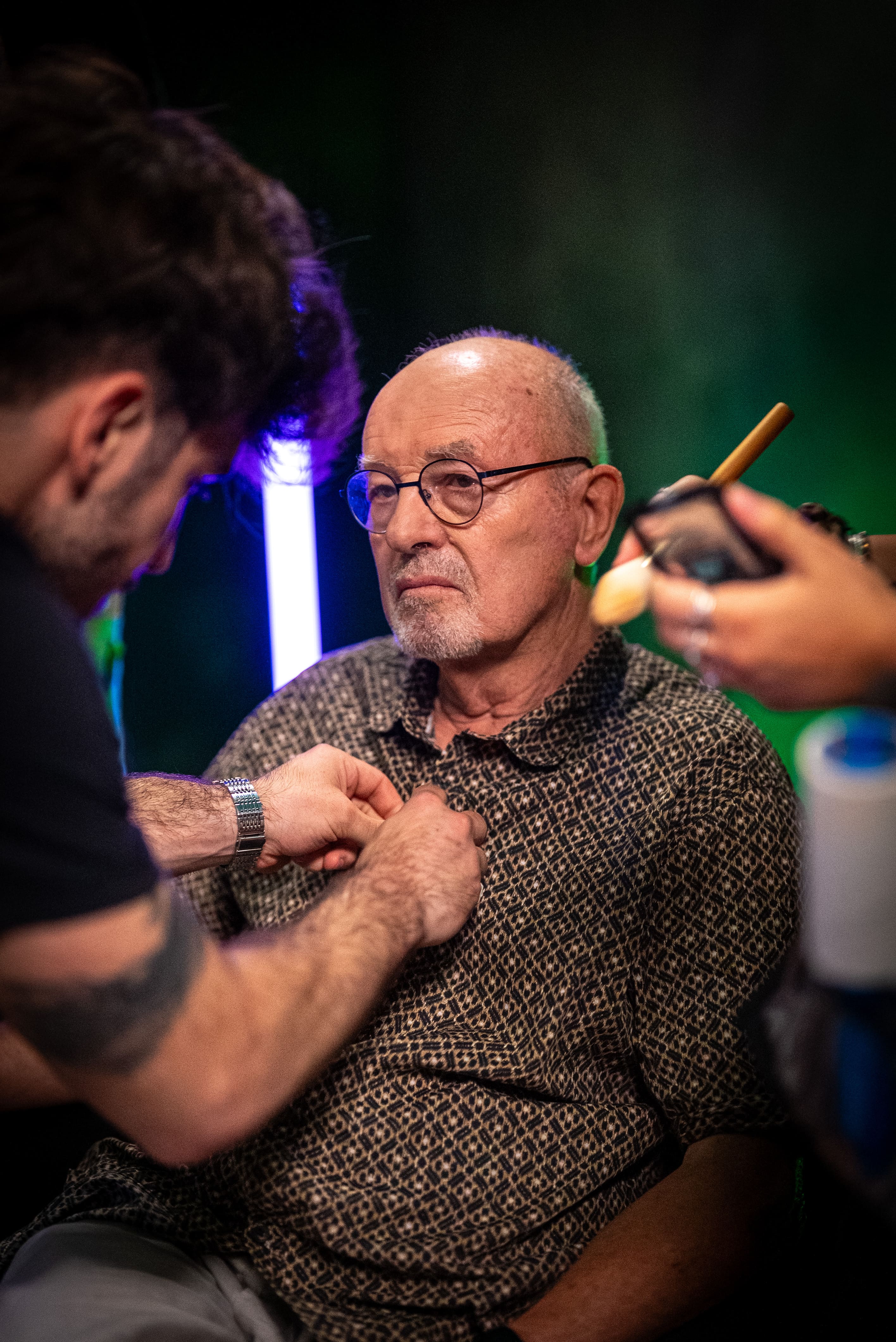 {   Photographie des coulisses (Behind the Scenes) dans le studio Legend, montrant un invité plus âgé, chauve, portant des lunettes rondes et une chemise à motifs sombres. Un membre de l'équipe (visible au premier plan) ajuste son col, tandis qu'une autre main (à droite) applique du maquillage ou de la poudre pour la caméra. L'arrière-plan présente des néons lumineux (vert et violet/bleu). Cette image illustre la préparation professionnelle avant le tournage d'une interview ou d'un podcast, soulignant l'expertise de Nux-Studio en production photo, direction artistique et gestion des coulisses pour les médias.