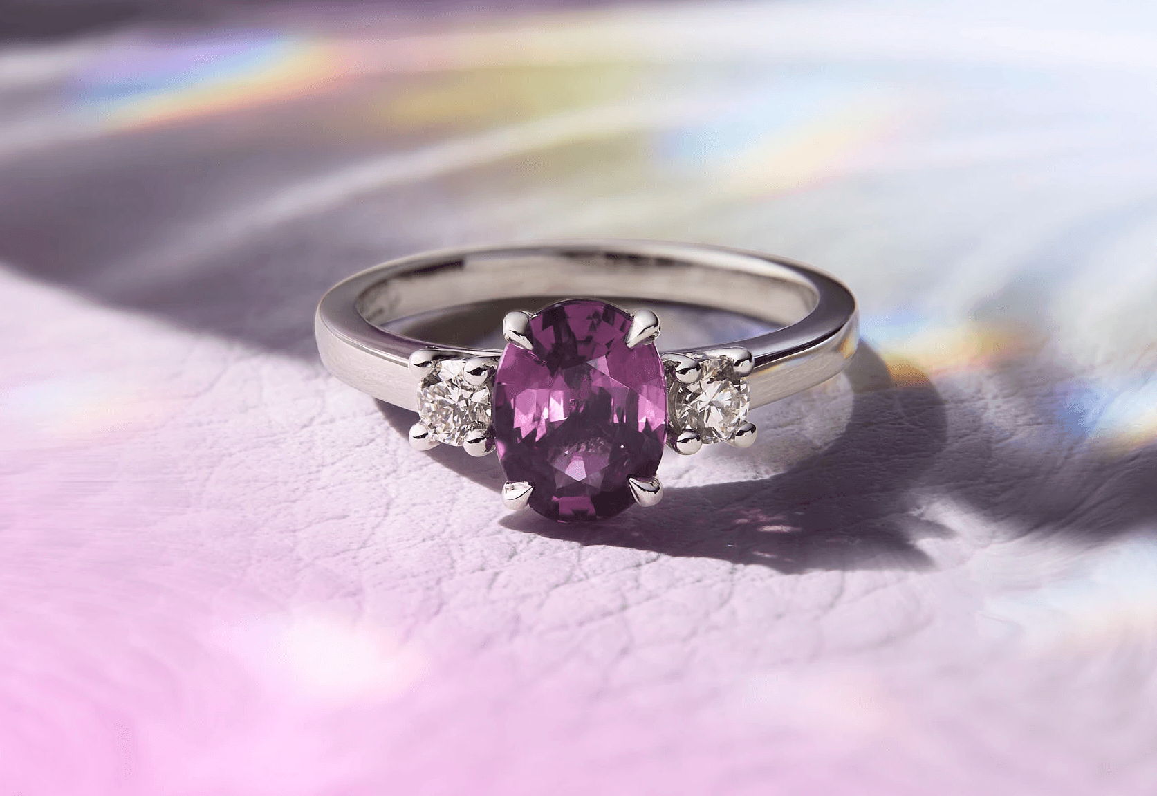A silver ring with a purple gemstone rests on a softly colored surface, reflecting light.