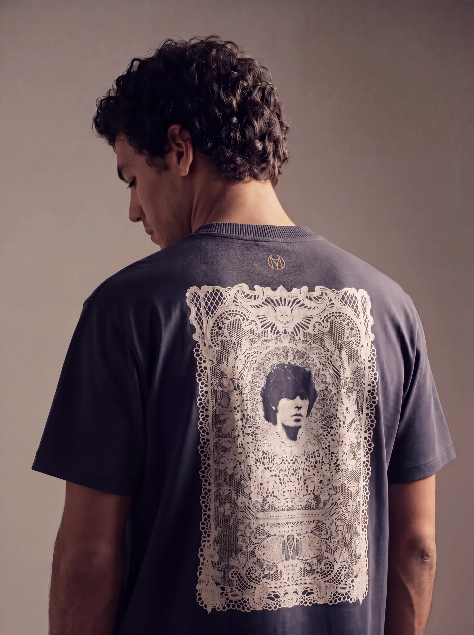 Back view of a male model wearing a dark grey Maradona Official t-shirt featuring an ornate lace-framed portrait graphic of Diego Maradona