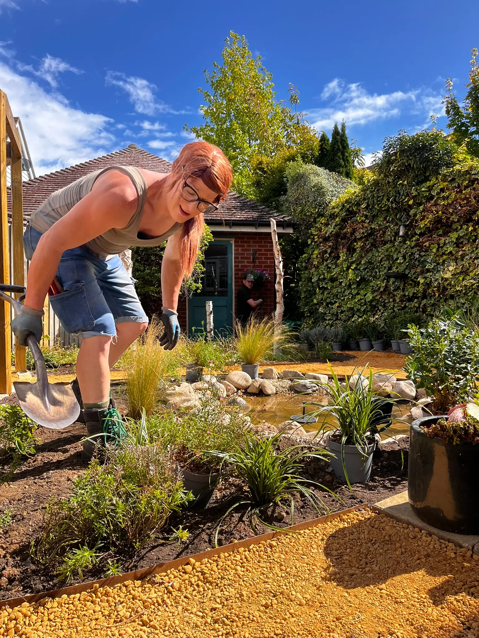 A person in shorts gardens in a sunny backyard surrounded by green plants and a blue sky.