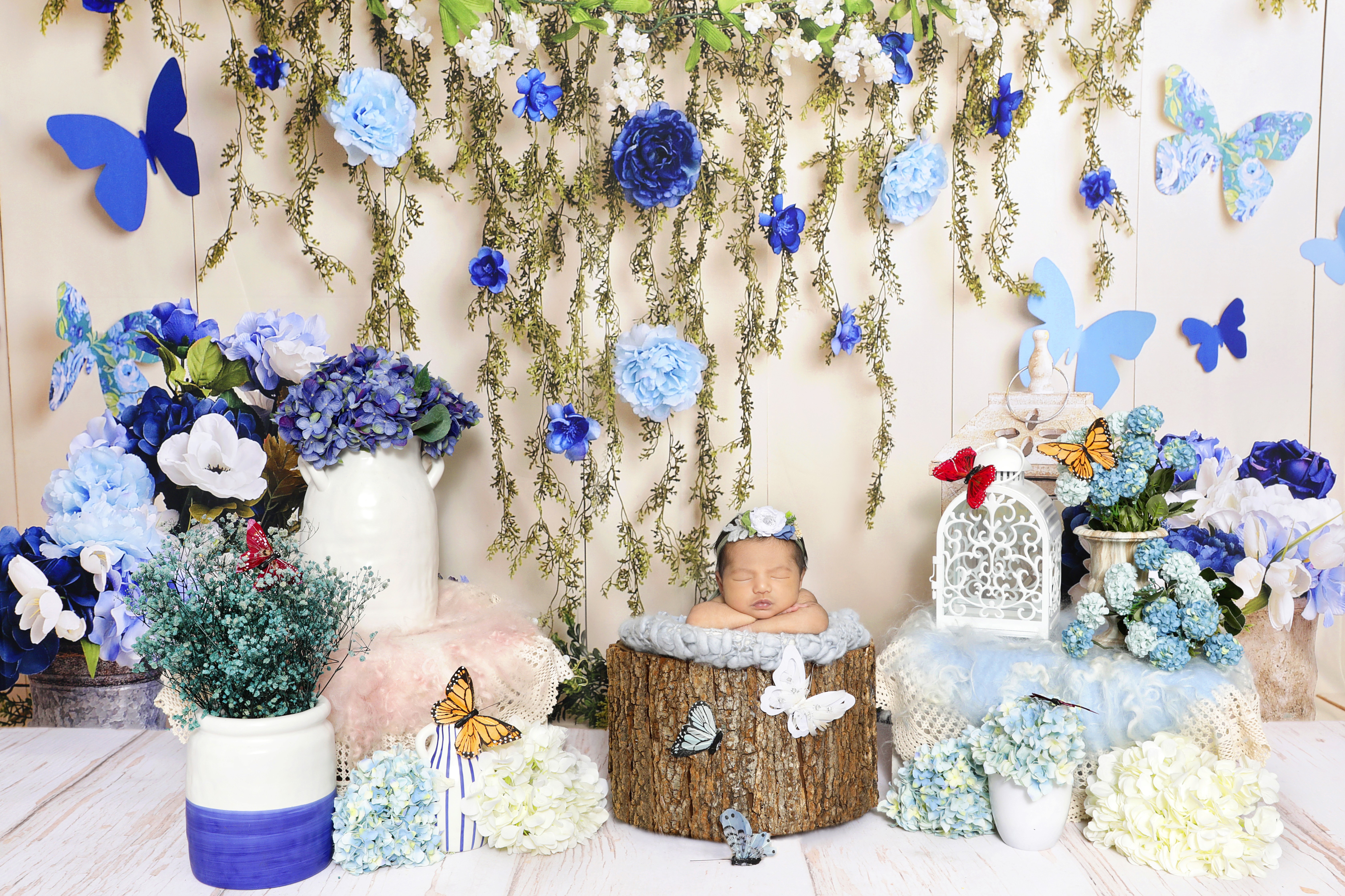 A newborn sleeps in a wooden bucket surrounded by blue flowers, hanging greenery, and butterfly decor in this infant photography setup.