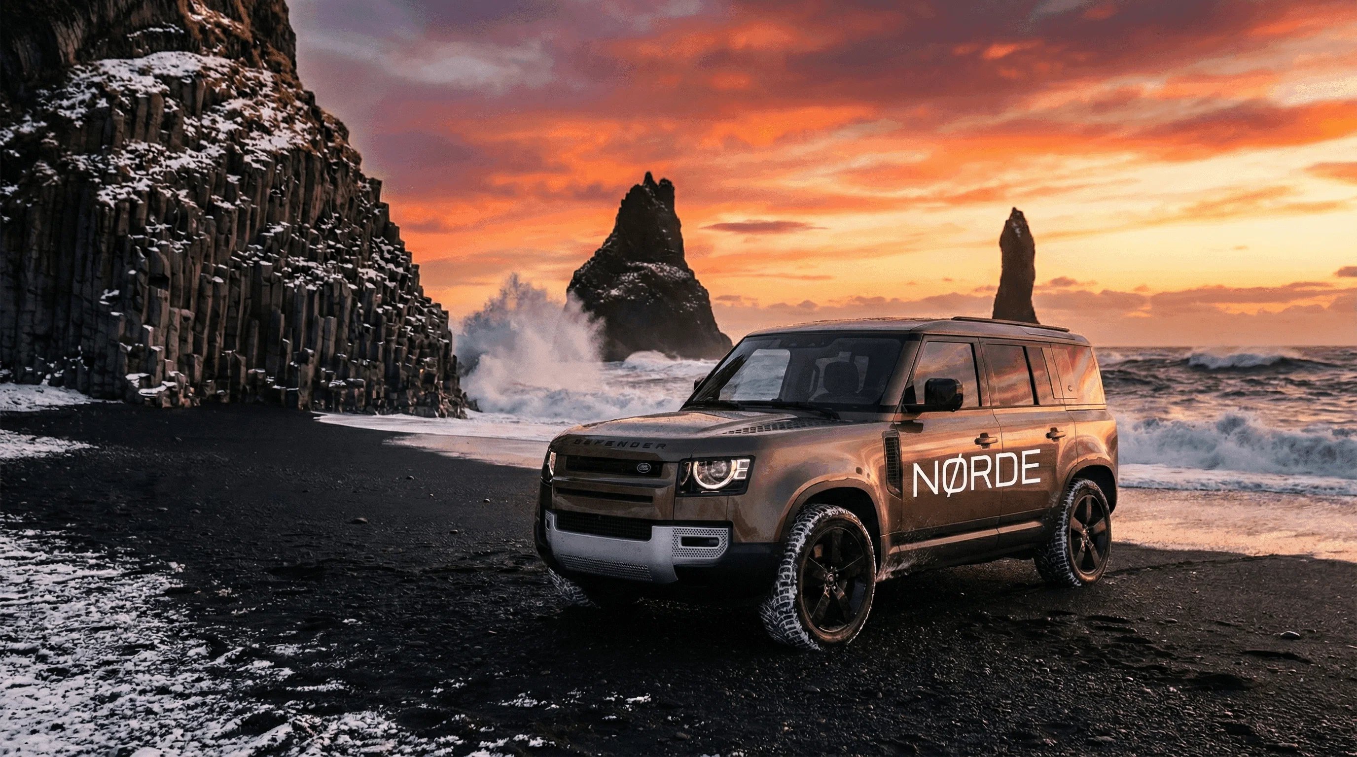 A brown Land Rover Defender parked on a black sand beach with sea stacks and a colorful sunset in the background.