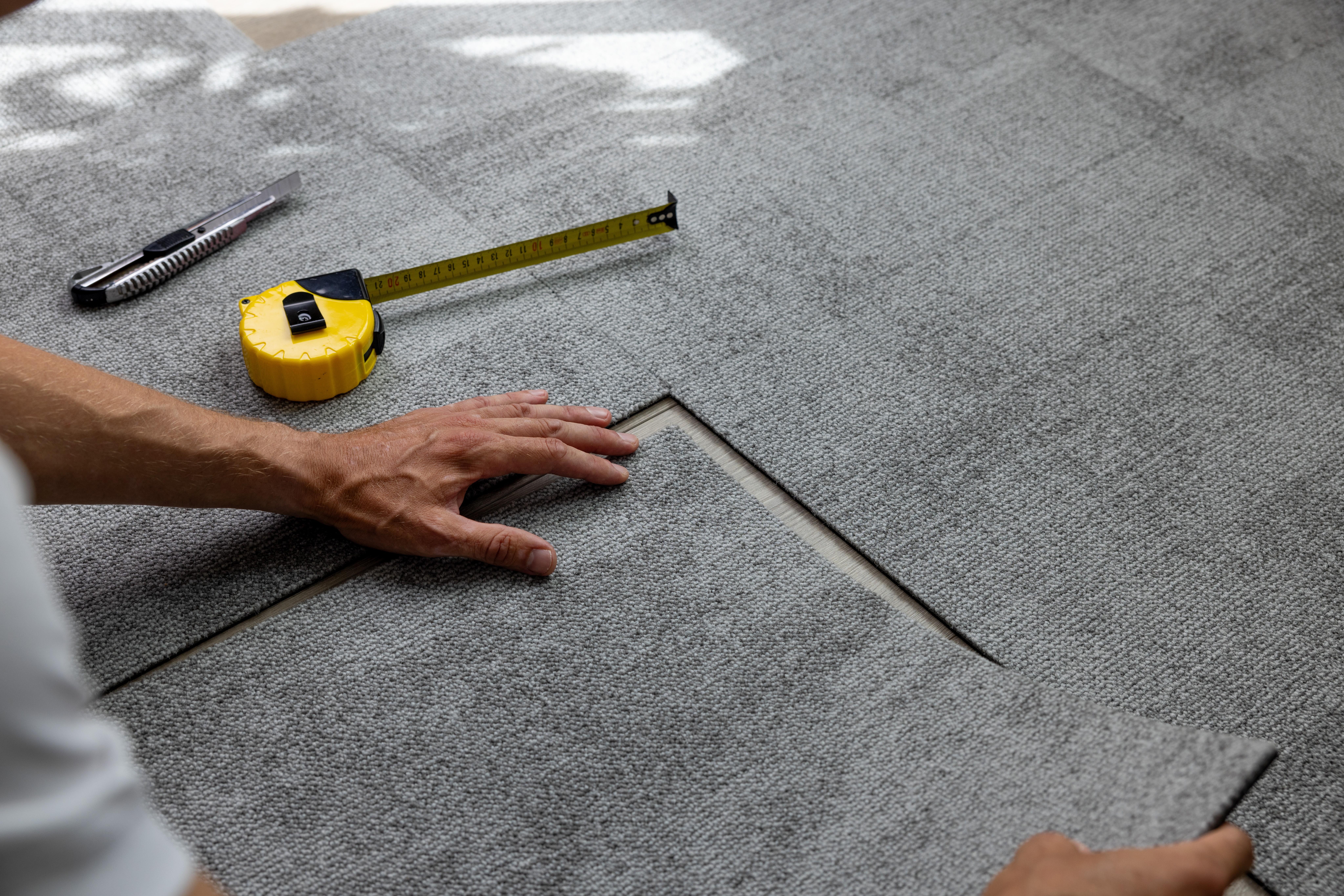 Installer setting a grey carpet tile using a tape measure and utility knife