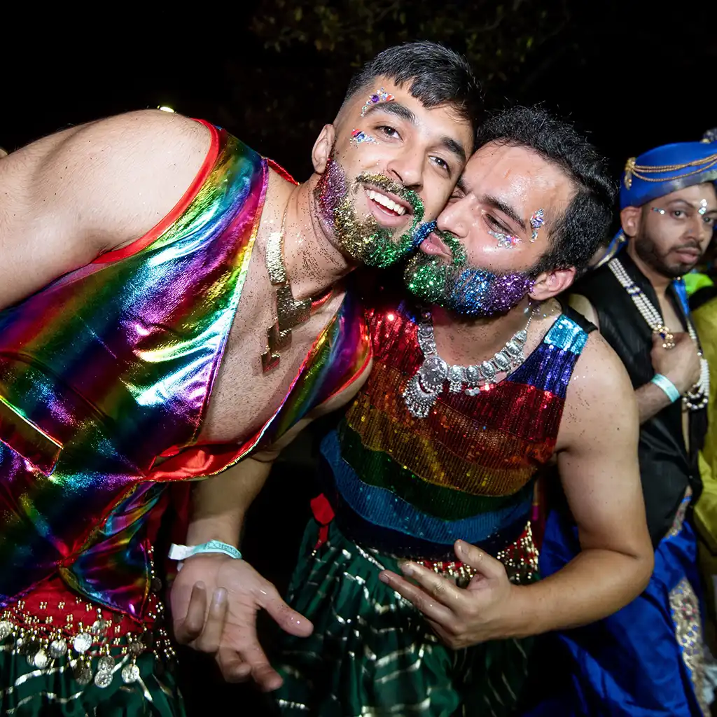 Two people in rainbow outfits and glittery beards laugh joyfully at a lively party. A third person in a colorful turban is seen in the background.
