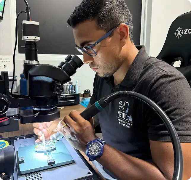 Especialista em microsoldagem da NETOCell trabalhando em placa eletrônica sob microscópio estéreo em bancada técnica iluminada com ferramentas e soprador de fumaça — reparo profissional de celulares e eletrônicos