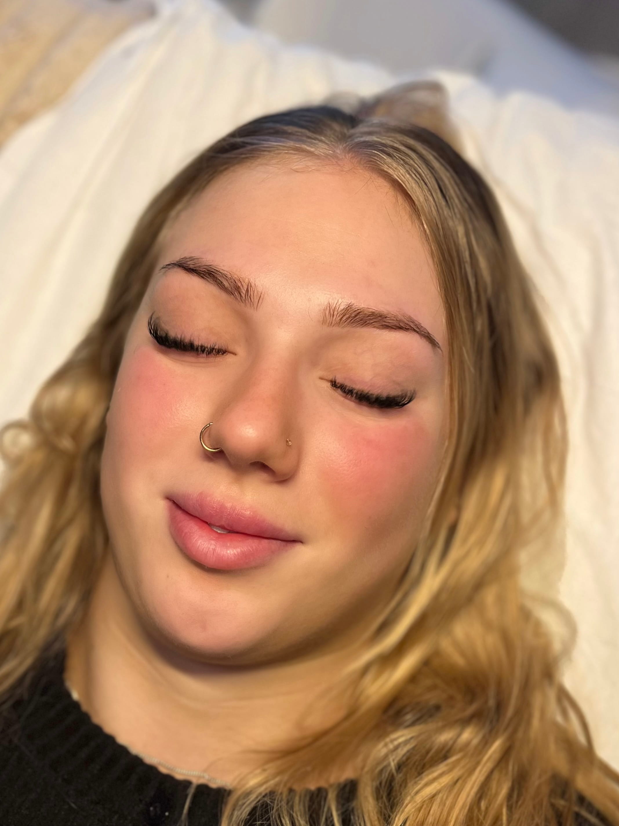 Woman with closed eyes, light red cheeks, and relaxed expression. She has long blonde hair and a nose ring, lying on a soft white pillow.