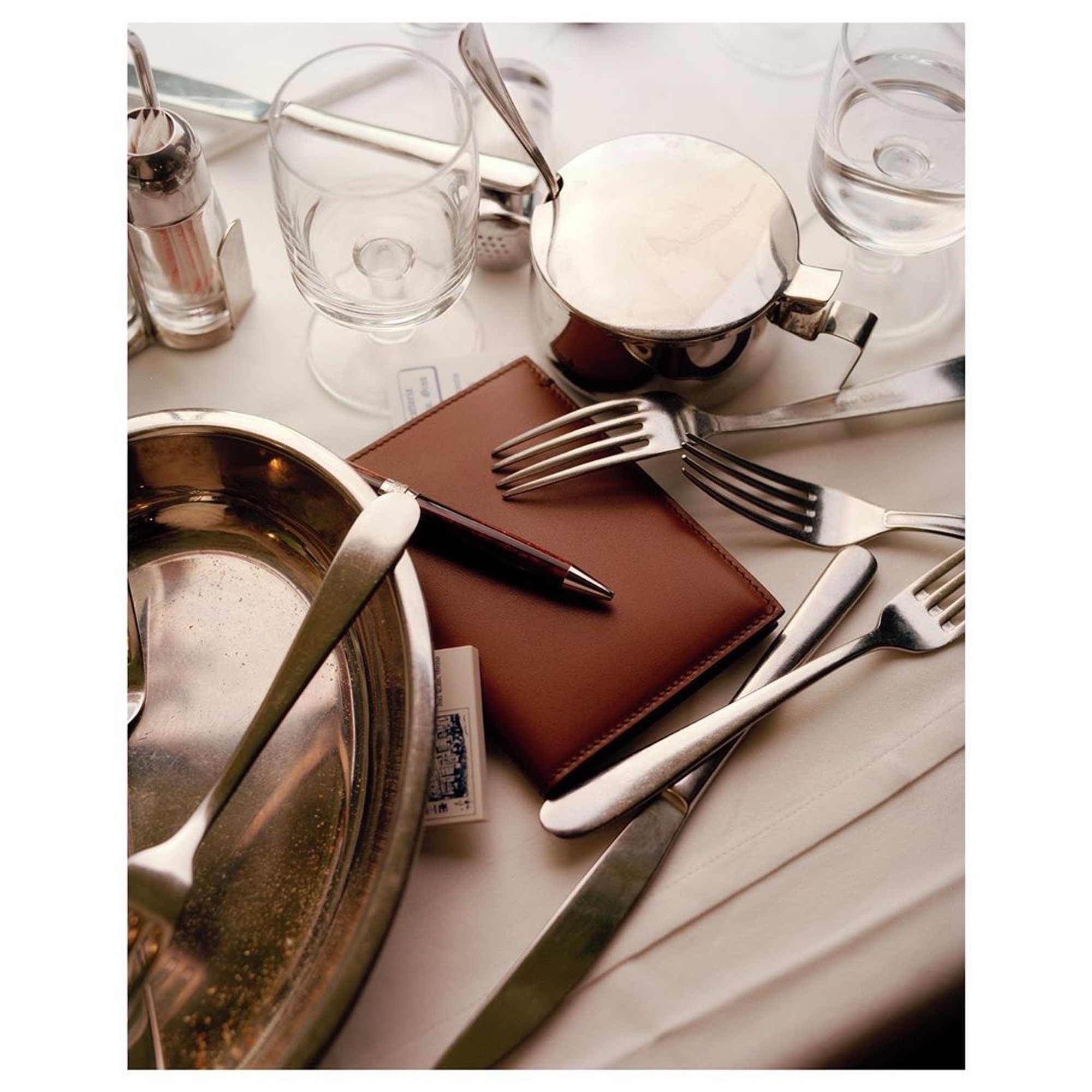 A brown leather notebook and pen lie among silver cutlery, an empty silver dish and glasses on a white tablecloth in a restaurant.
