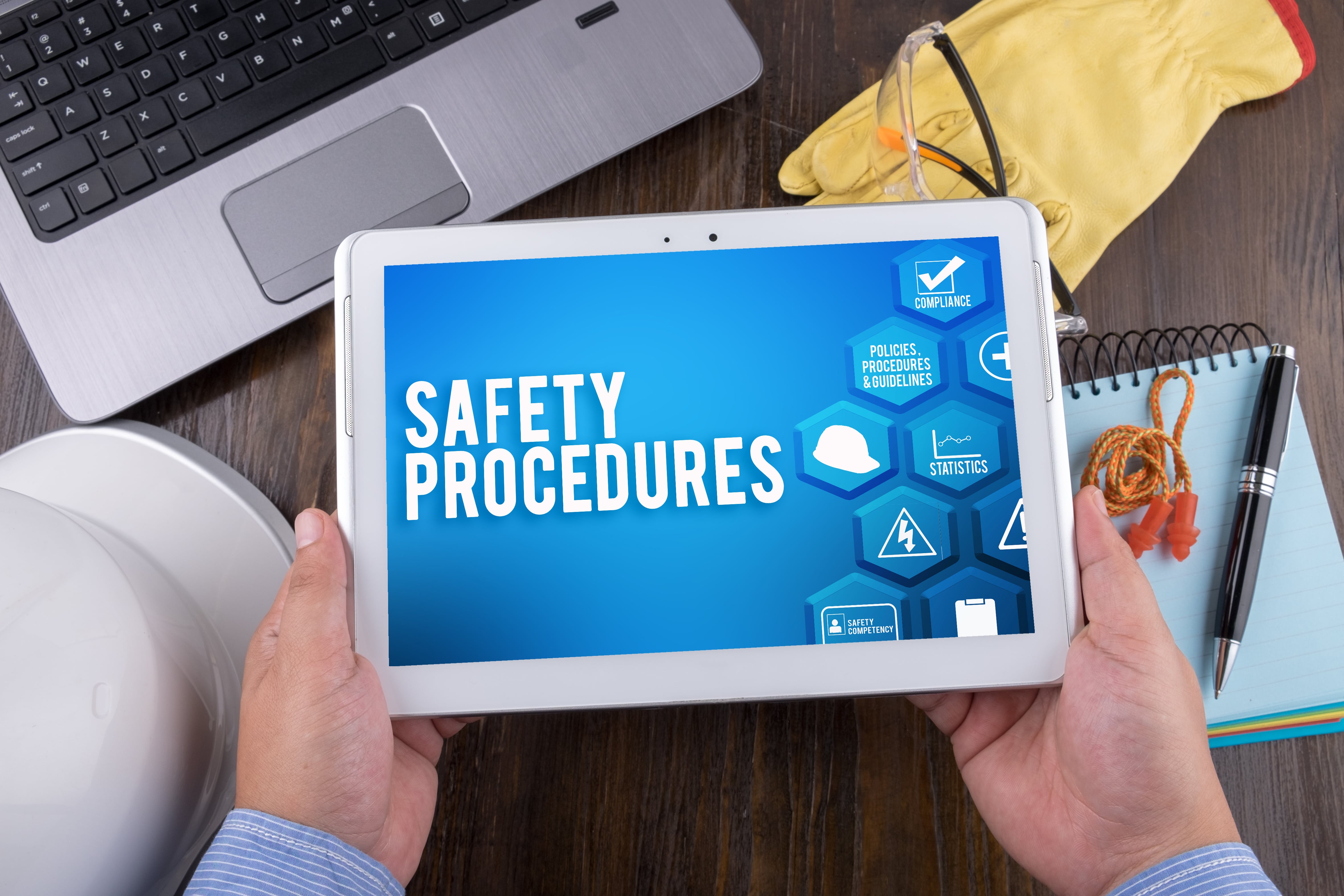 A person holding a tablet displaying “Safety Procedures,” with safety gear, a laptop, and a notepad on a desk.