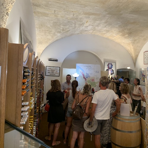 กลุ่มคนในร้านไวน์ที่มีเพดานโค้ง กำลังเดินเลือกซื้อและชิมไวน์ใกล้ชั้นวางและโต๊ะถังไวน์