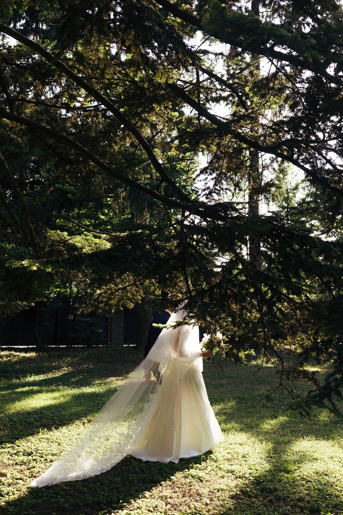 Sposa in abito bianco con strascico sotto albero secolare, luce naturale filtra tra le foglie nel giardino della villa