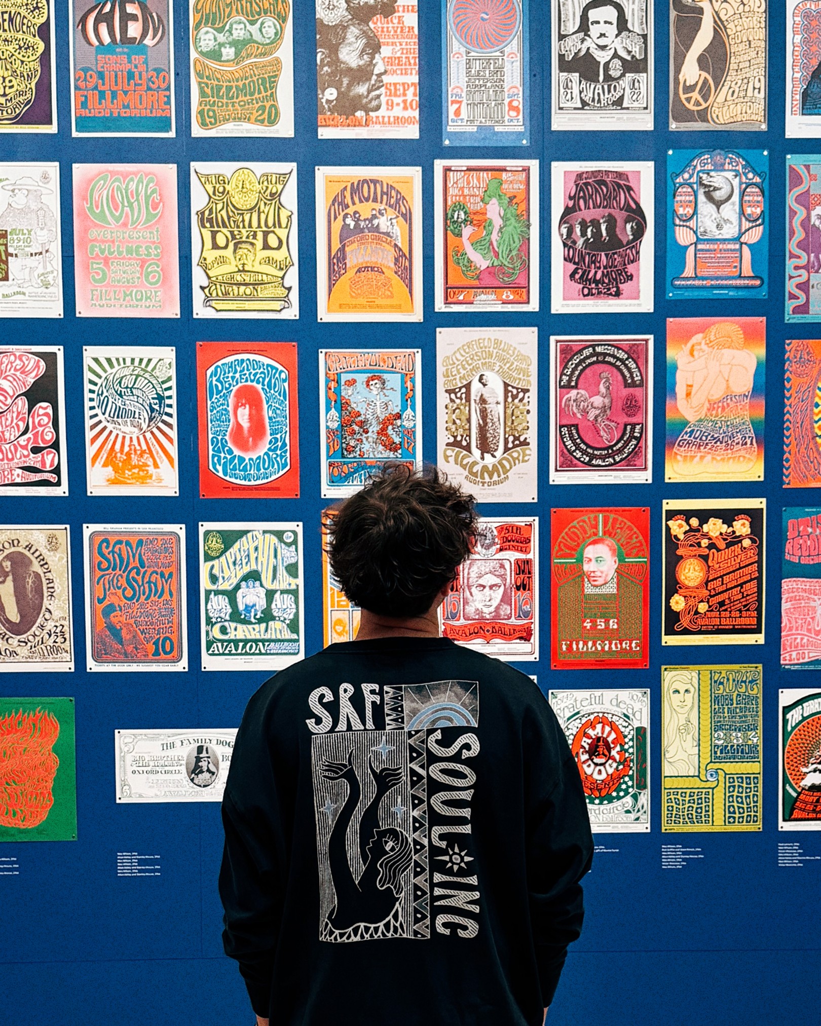 A person wearing a black sweatshirt with a graphic design on the back stands in front of a vibrant wall filled with vintage psychedelic music posters at the SFMOMA Art of Noise exhibition. The posters feature bold typography, intricate illustrations, and bright colors, showcasing the history of experimental music from the avant-garde to club culture.