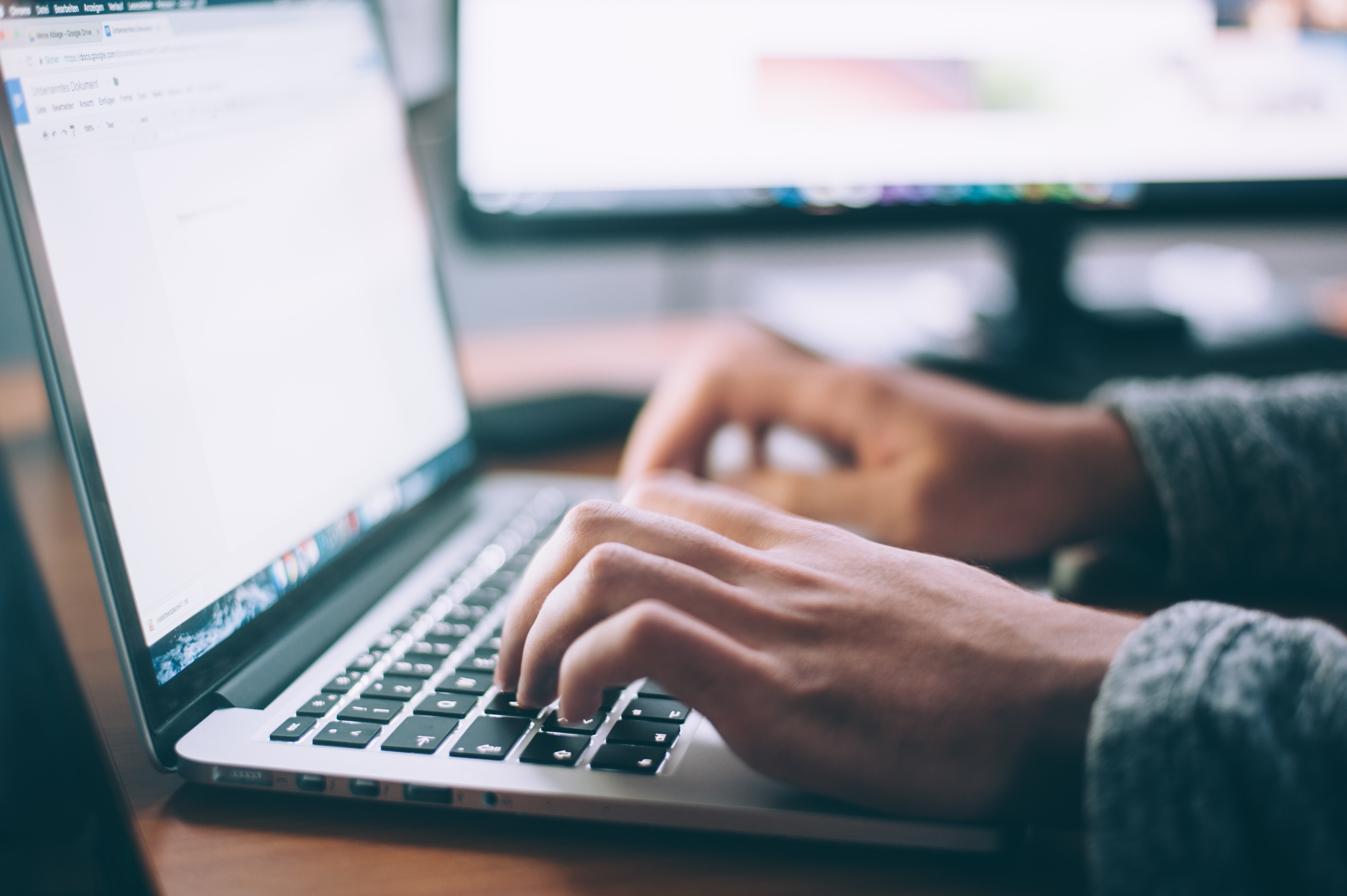 A person working on a laptop with a desktop screen next to the laptop