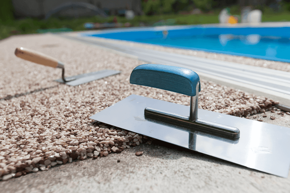 Two metal trowels, one with a blue handle and another with a wooden handle, resting on a textured concrete surface near a swimming pool. The tools suggest ongoing or completed poolside construction work.