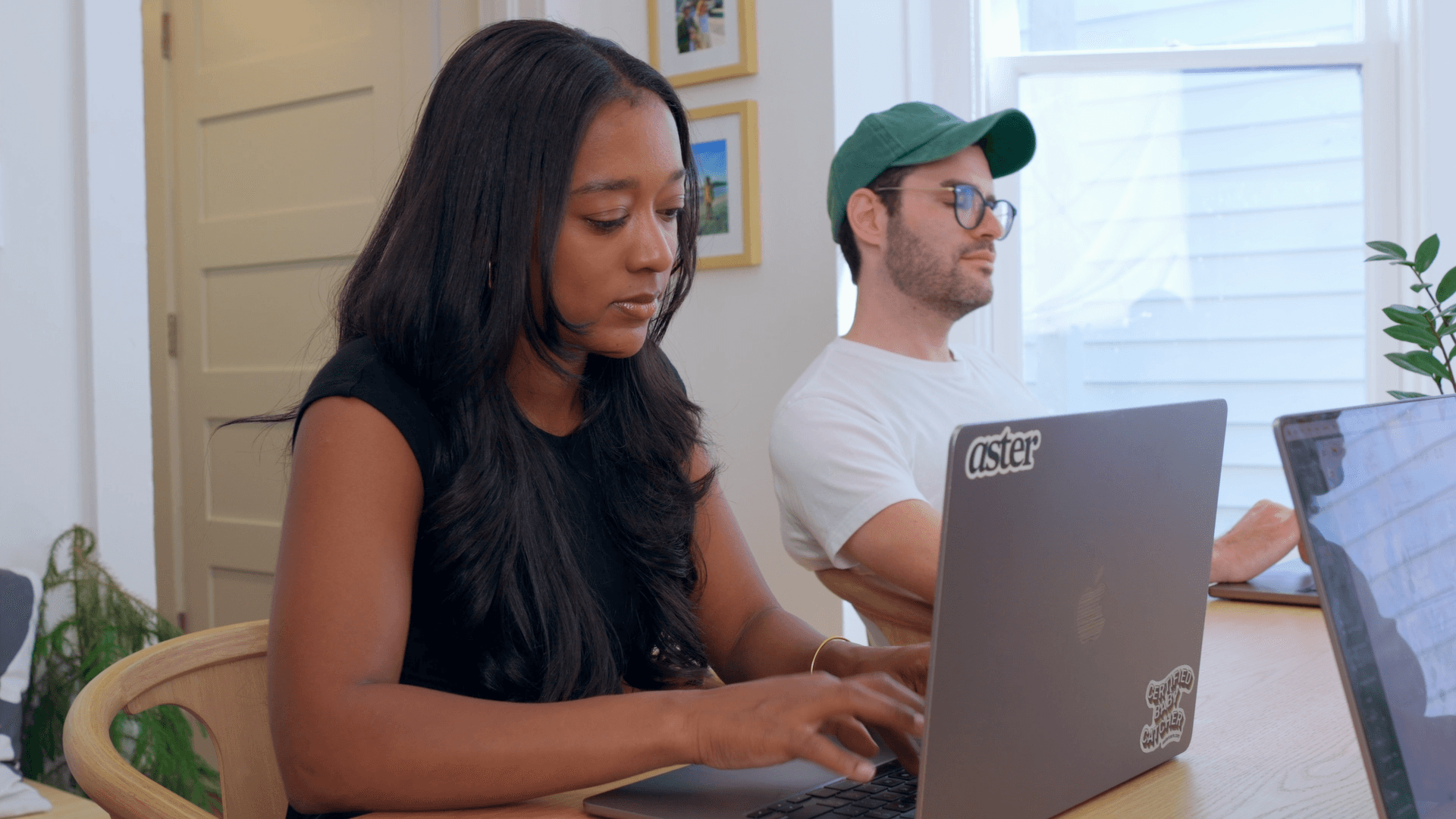 A smiling man and woman sit together on a sofa in a sunlit room, with large green potted plants in the background, embodying a warm and cheerful atmosphere.