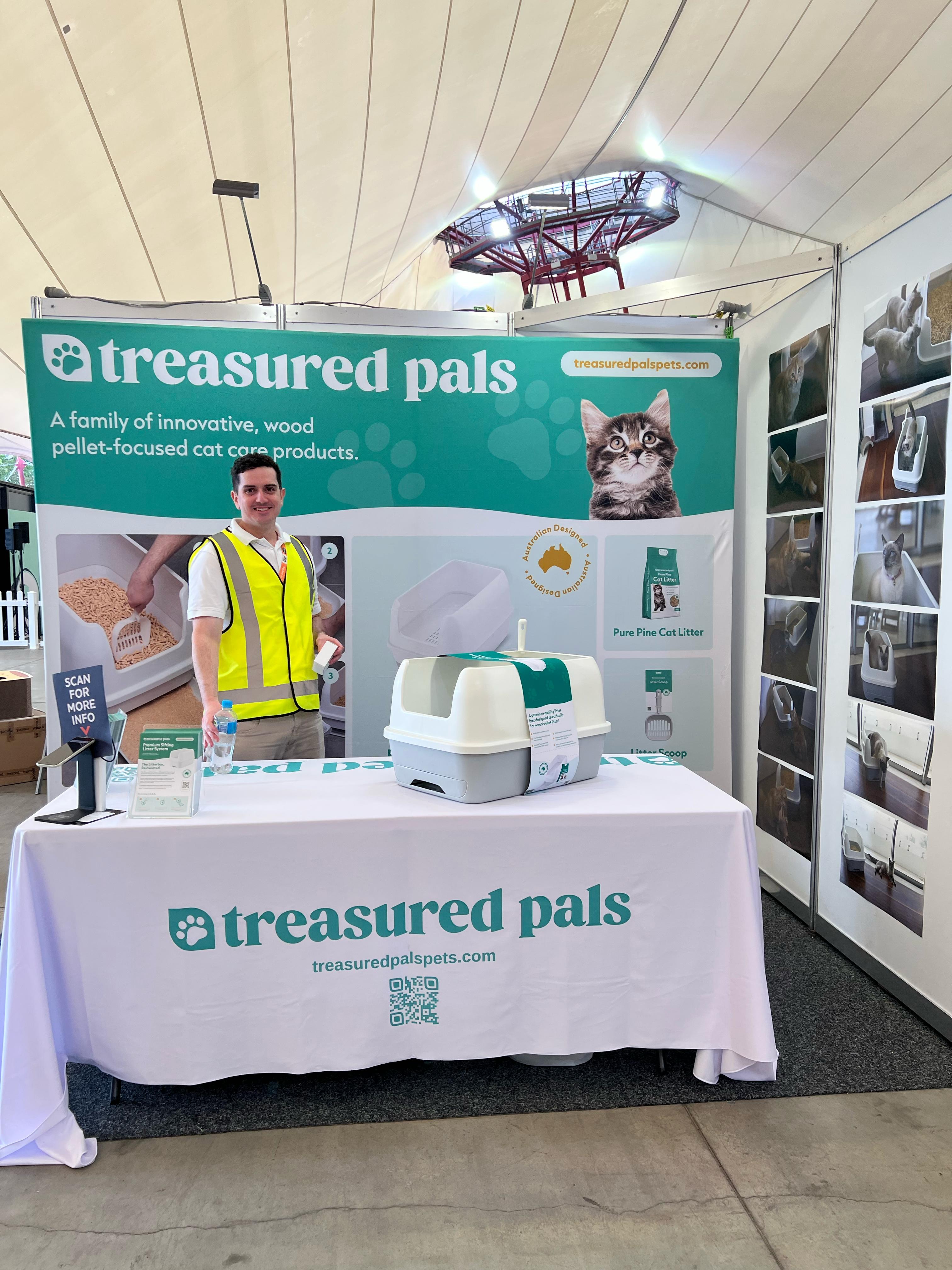 A fluffy, black and white cat sits inside a A Treasured Pals Premium Sifting Litter System  on a wooden floor, with a colorful striped object in the background.