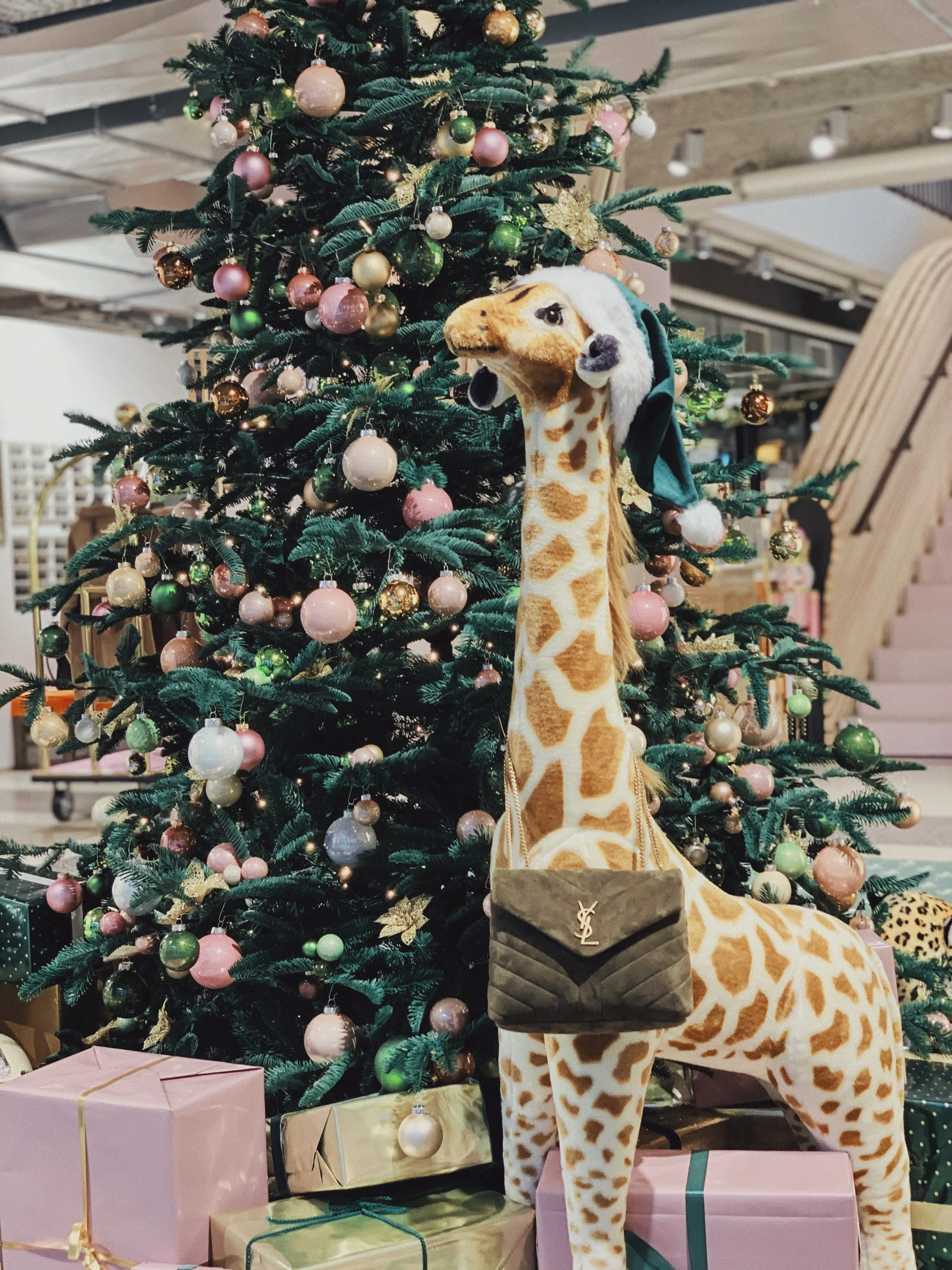 Plush giraffe with Santa hat carrying a green designer handbag in front of a Christmas tree with pink, green and gold baubles and pastel gift boxes at UZWEI.