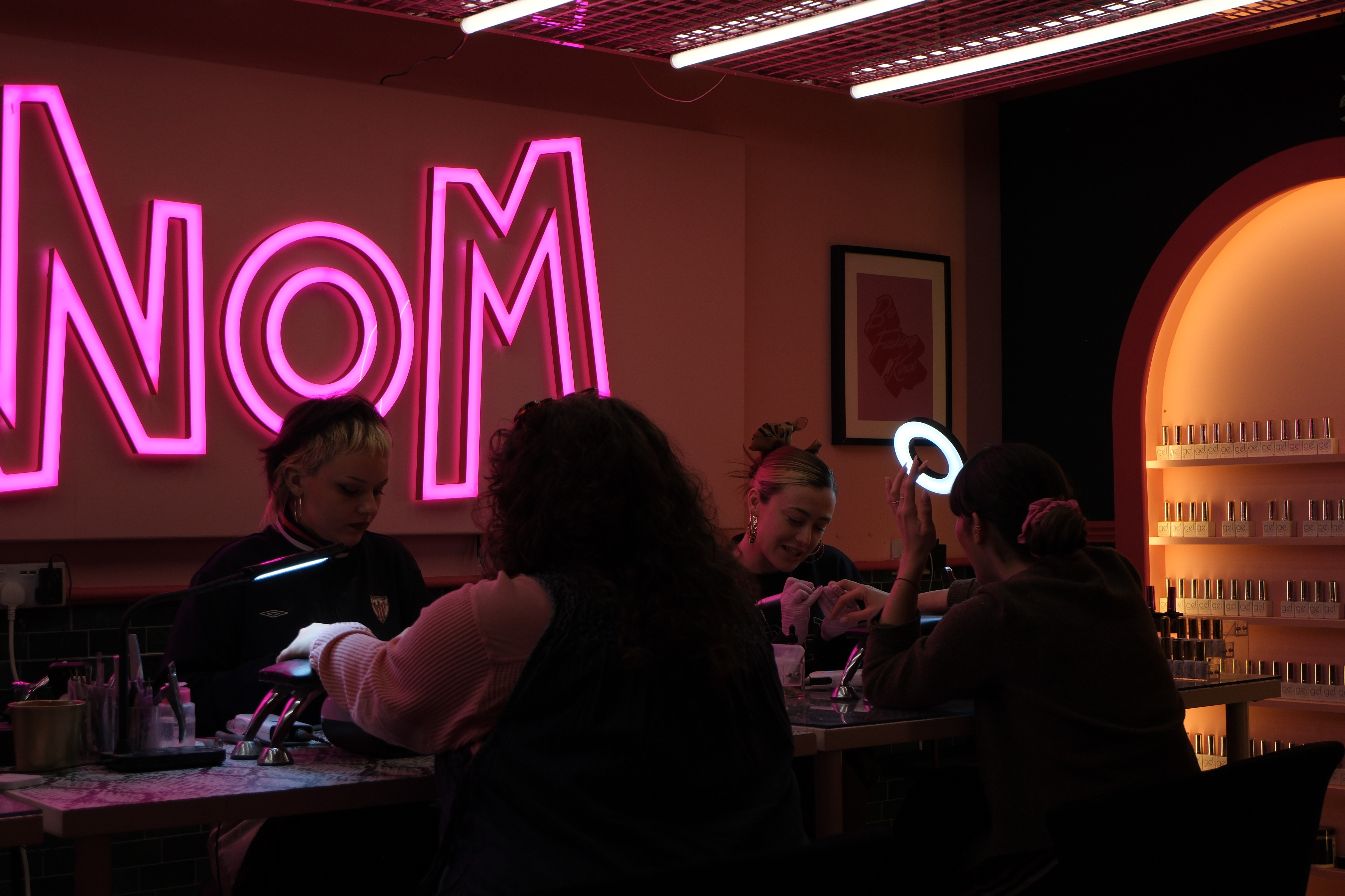 A wide, atmospheric shot of the salon during a busy session. Two nail technicians are at work with their clients at a snakeskin-patterned desk. The scene is dominated by a large, glowing pink neon "VENOM" sign, with warm desk lamps providing task lighting in the moody, dark-toned room.