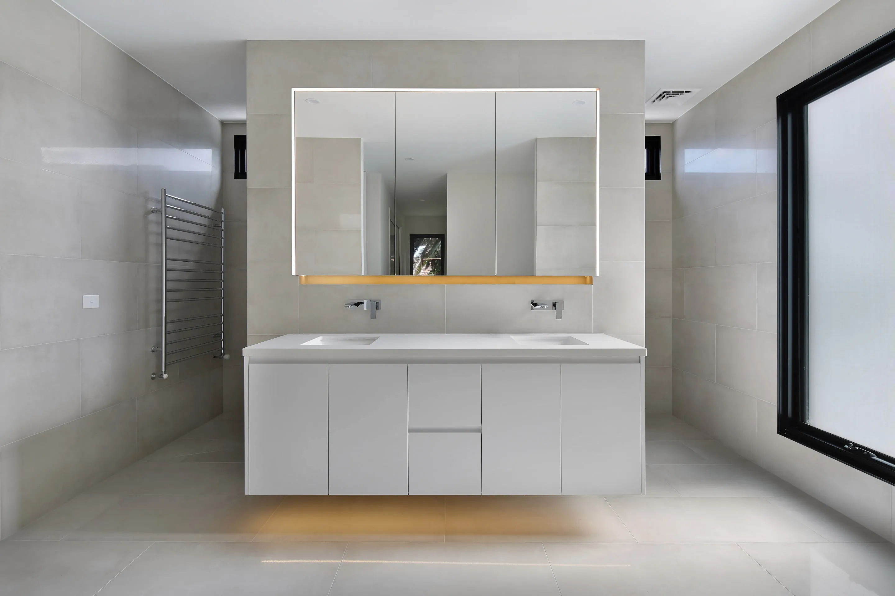 Bathroom in beige stone-look tiles with a white double vanity, mirrored cabinet with under-shelf lighting, chrome wall-mounted taps, a heated towel rail and a frosted black-framed window