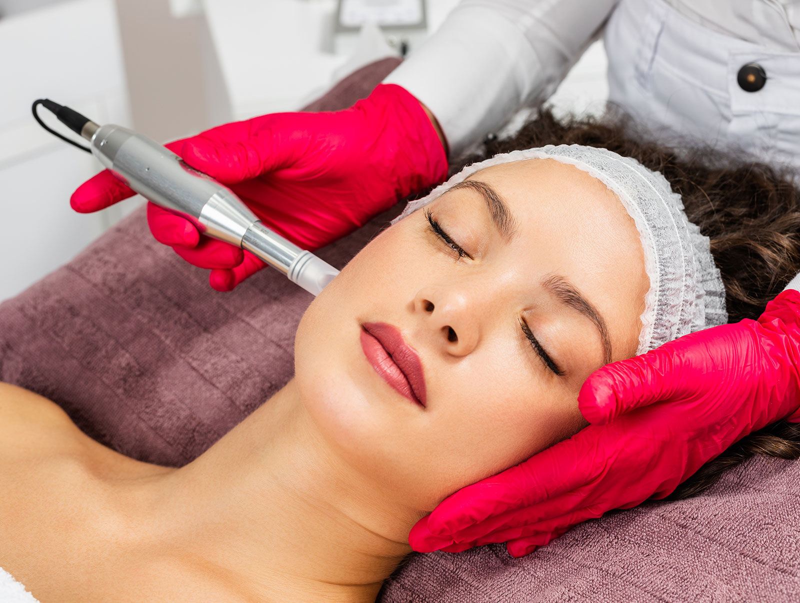 &nbsp;A woman receiving a microneedling treatment on her cheek by a professional wearing red gloves, with a specialized device.
