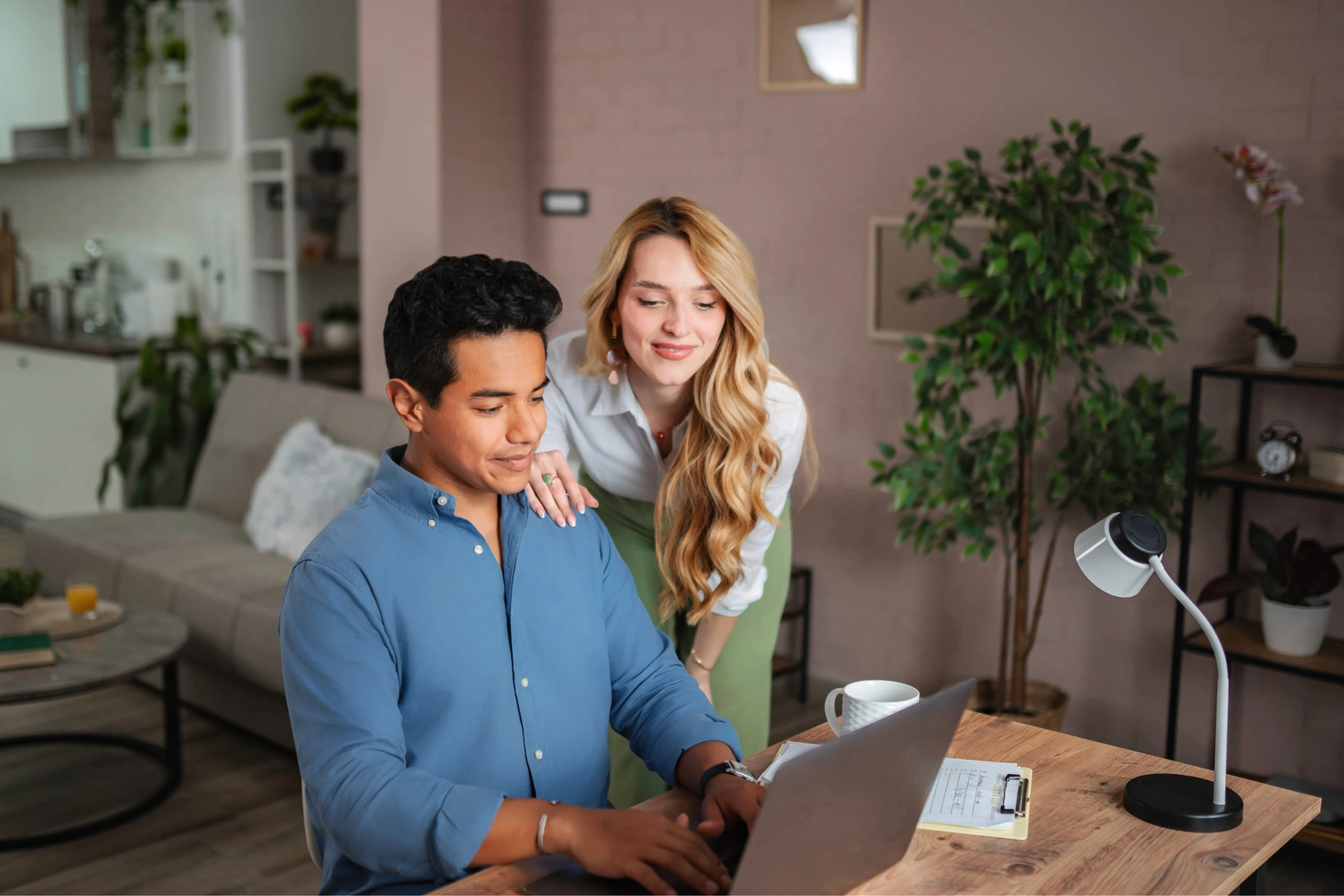 A man in a blue shirt sits at a wooden desk using a laptop, while a woman in a white blouse leans over his shoulder in a modern living room with plants and a cozy kitchen in the background.