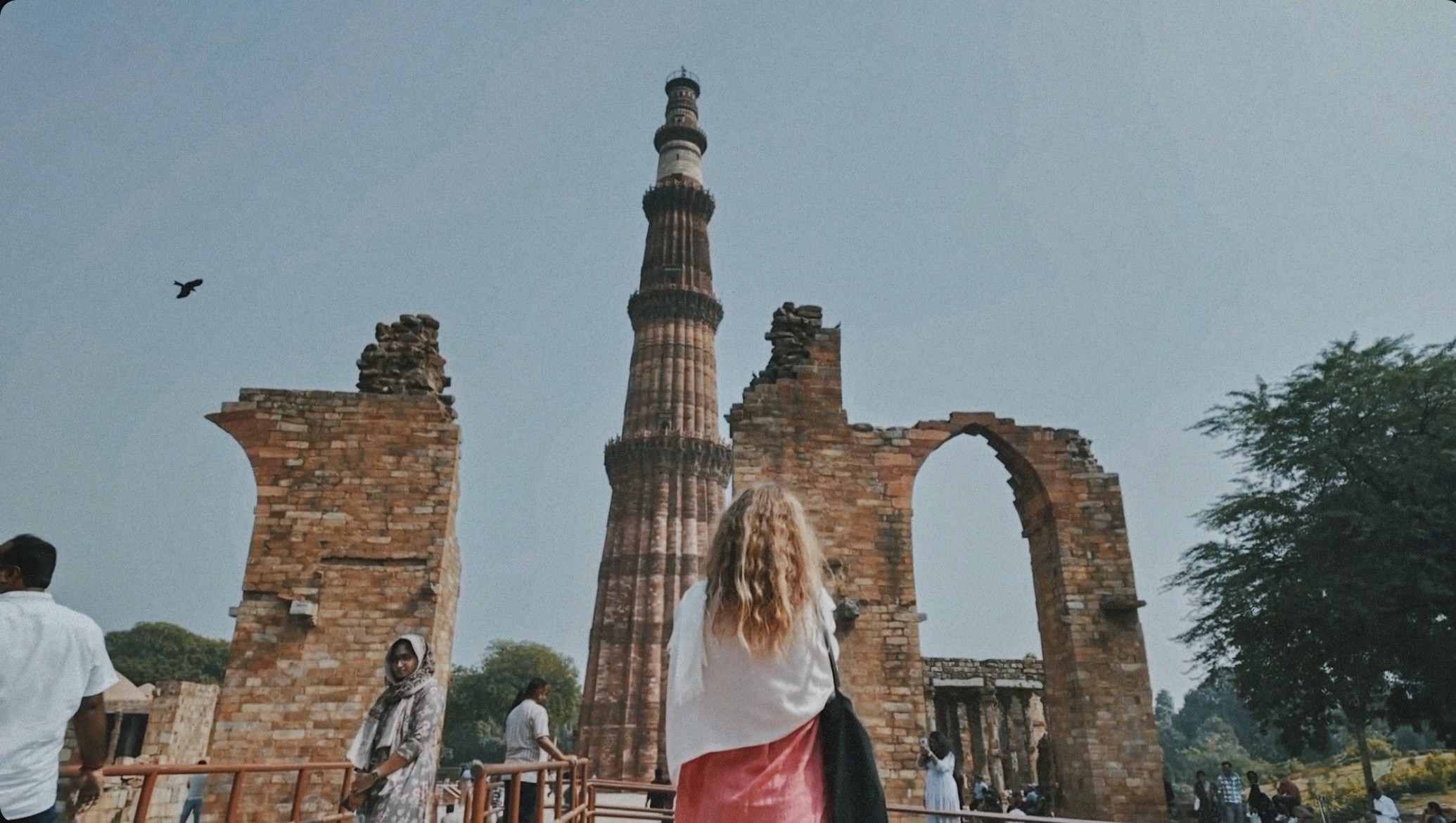 Back view of a woman admiring the Qutub Minar in Delhi, surrounded by lush greenery and historic architecture. Explore India's rich heritage with a journey from Delhi's iconic landmarks to the serene hills of Dharamshala. Ideal for those seeking a blend of cultural discovery and spiritual retreat.