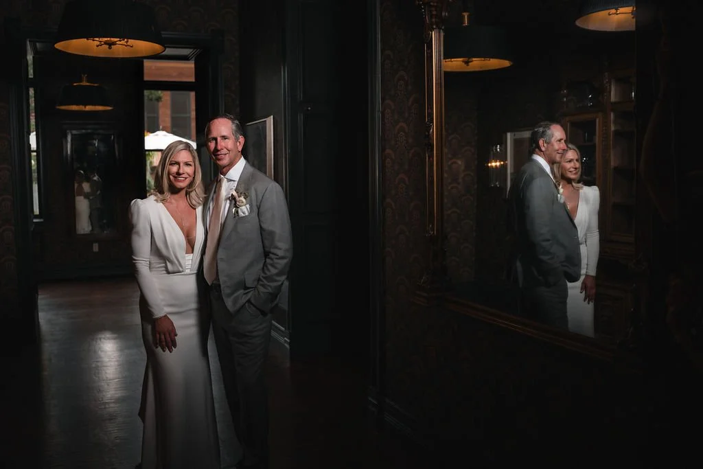 A bride and groom pose beside a large mirror that reflects them in a dimly lit hallway.