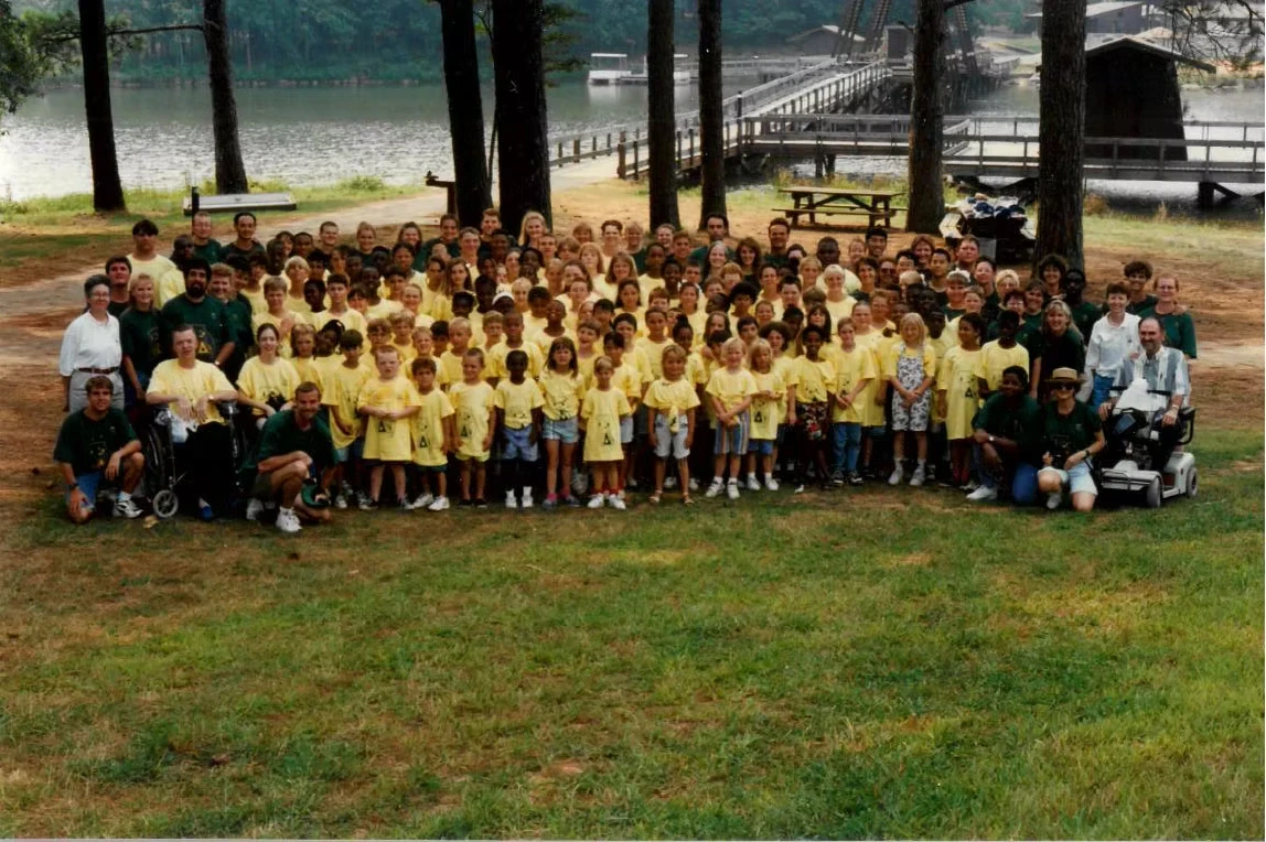 Large group photo of campers and staff wearing yellow Camp Juliena shirts, posing outdoors near a lake and wooden bridge.
