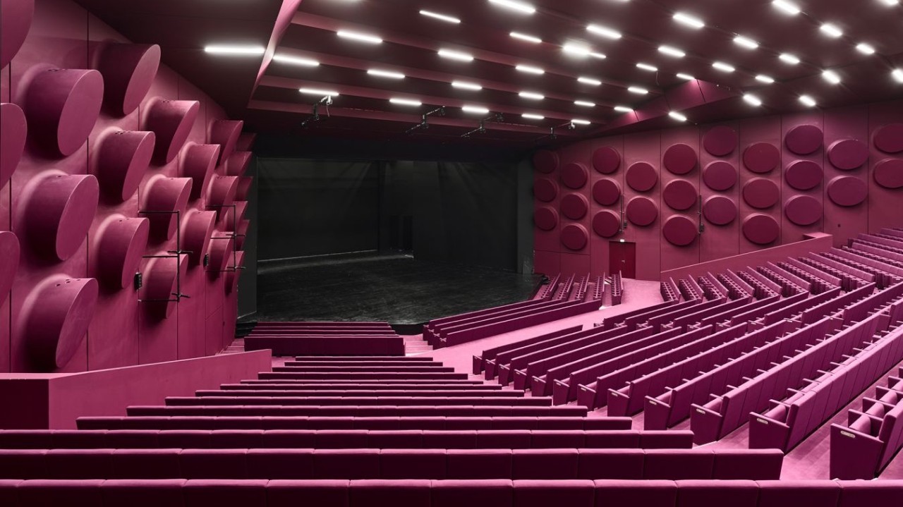 Vue d’un auditorium aux rangées de sièges violets orientées vers une scène noire, sous un plafond éclairé de lignes lumineuses.