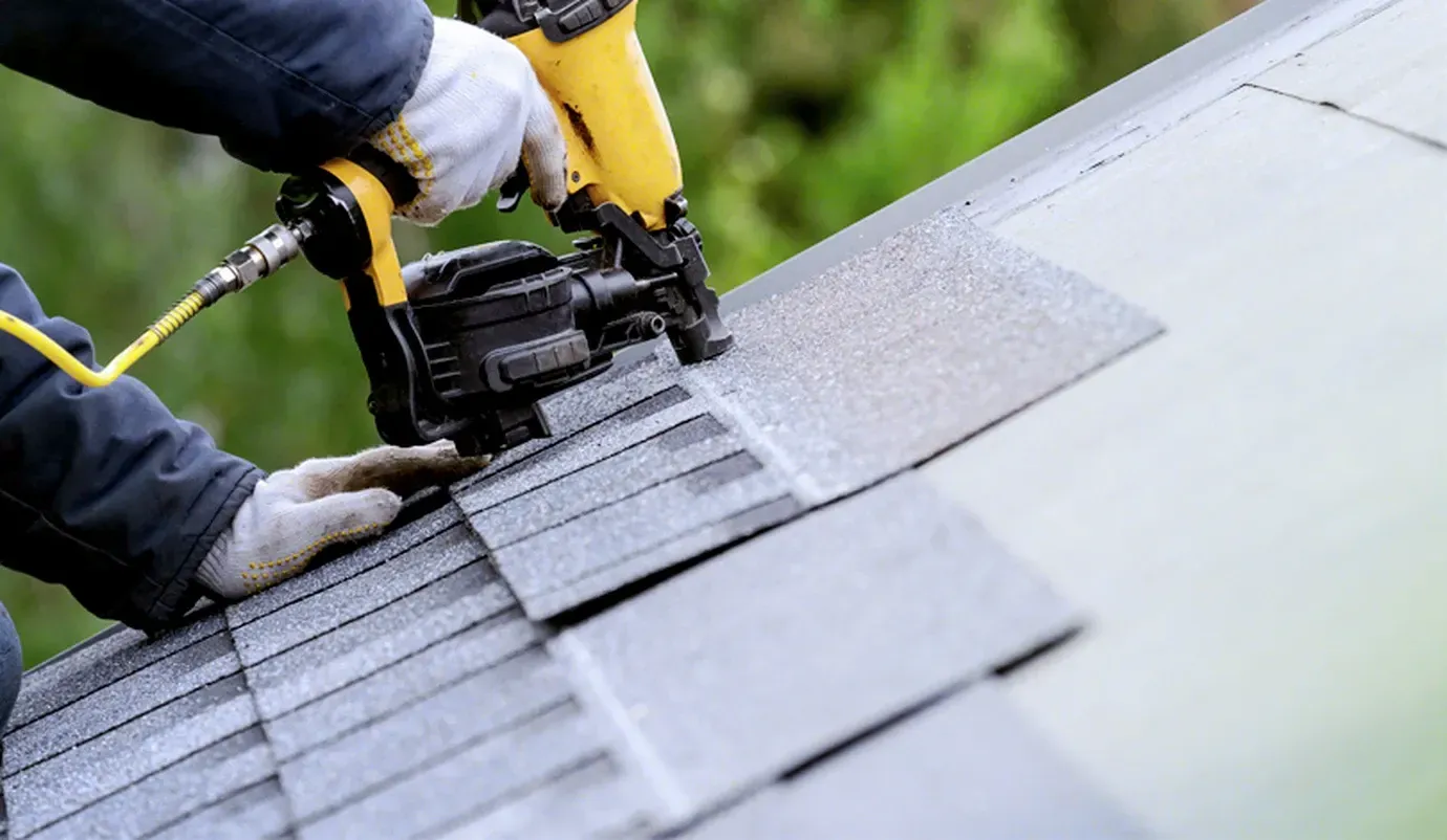 A person wearing gloves uses a yellow pneumatic nail gun to attach dark gray asphalt shingles to a sloped roof.