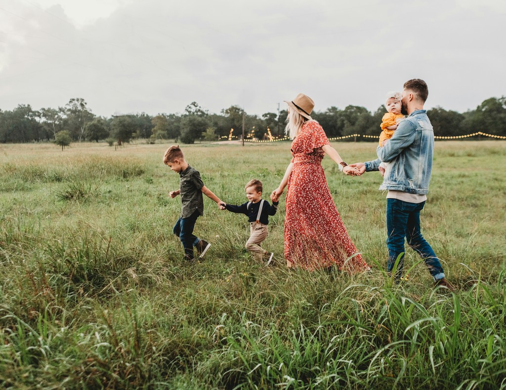 A cheerful family walking hand-in-hand through a grassy field, with the mother in a red dress and hat, the father holding a baby, and two young children leading the way, enjoying an outdoor adventure.