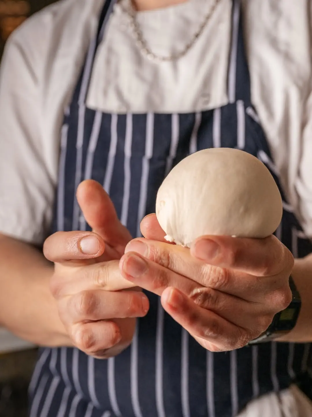 A chef wearing a black cap and white jacket with a red stripe sprinkles cheese over a freshly baked pizza, showcasing Italian culinary craftsmanship at a fine dining Italian restaurant in London.