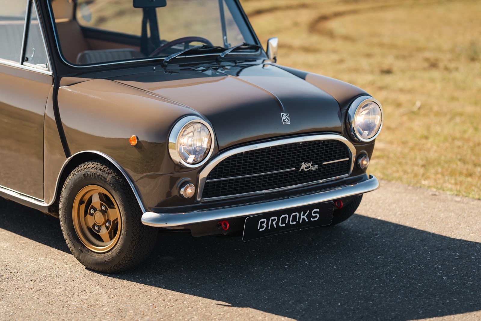 A close-up view of a vintage car with a rounded front, parked on a road with a grassy backdrop.