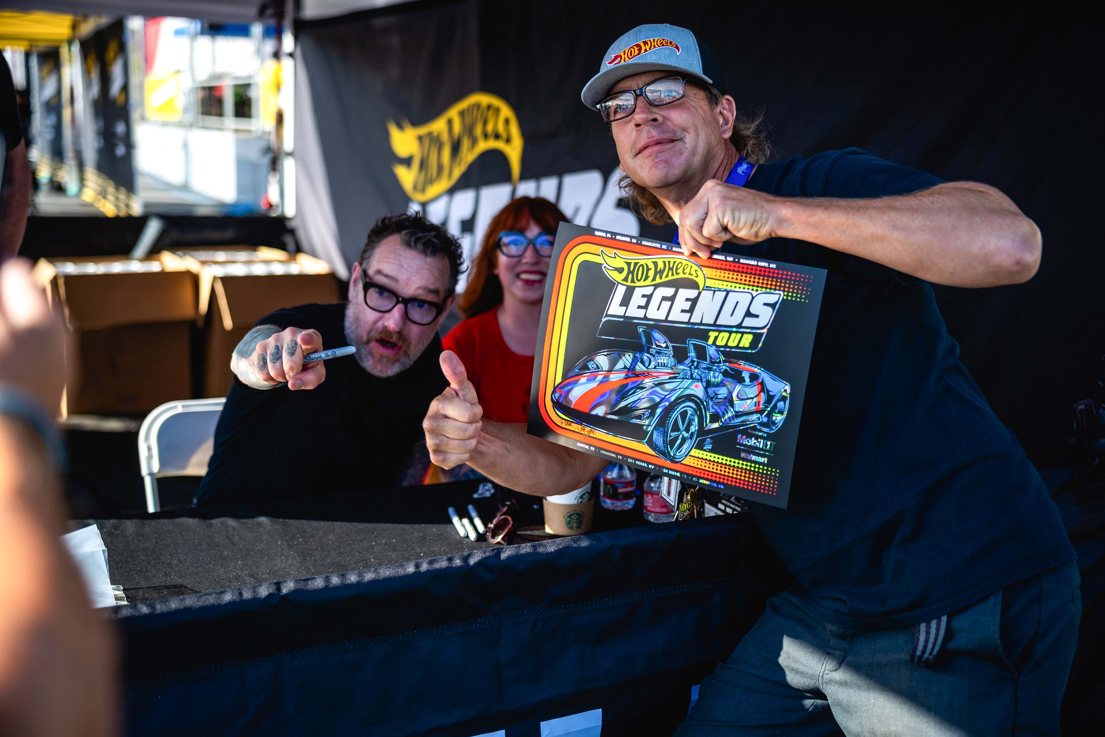 Artist Dirty Donny Gillies at a Hot Wheels Legends Tour event sitting at a signing table while a fan in a Hot Wheels cap holds up a colorful poster of the Mobil 1–branded Twin Mill car, both smiling and gesturing enthusiastically.