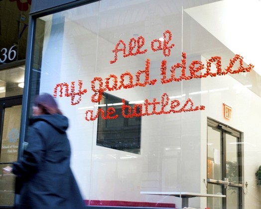 A photo of some orange pieces of plastic on a glass wall forming a text that says: All of my good ideas are battles.