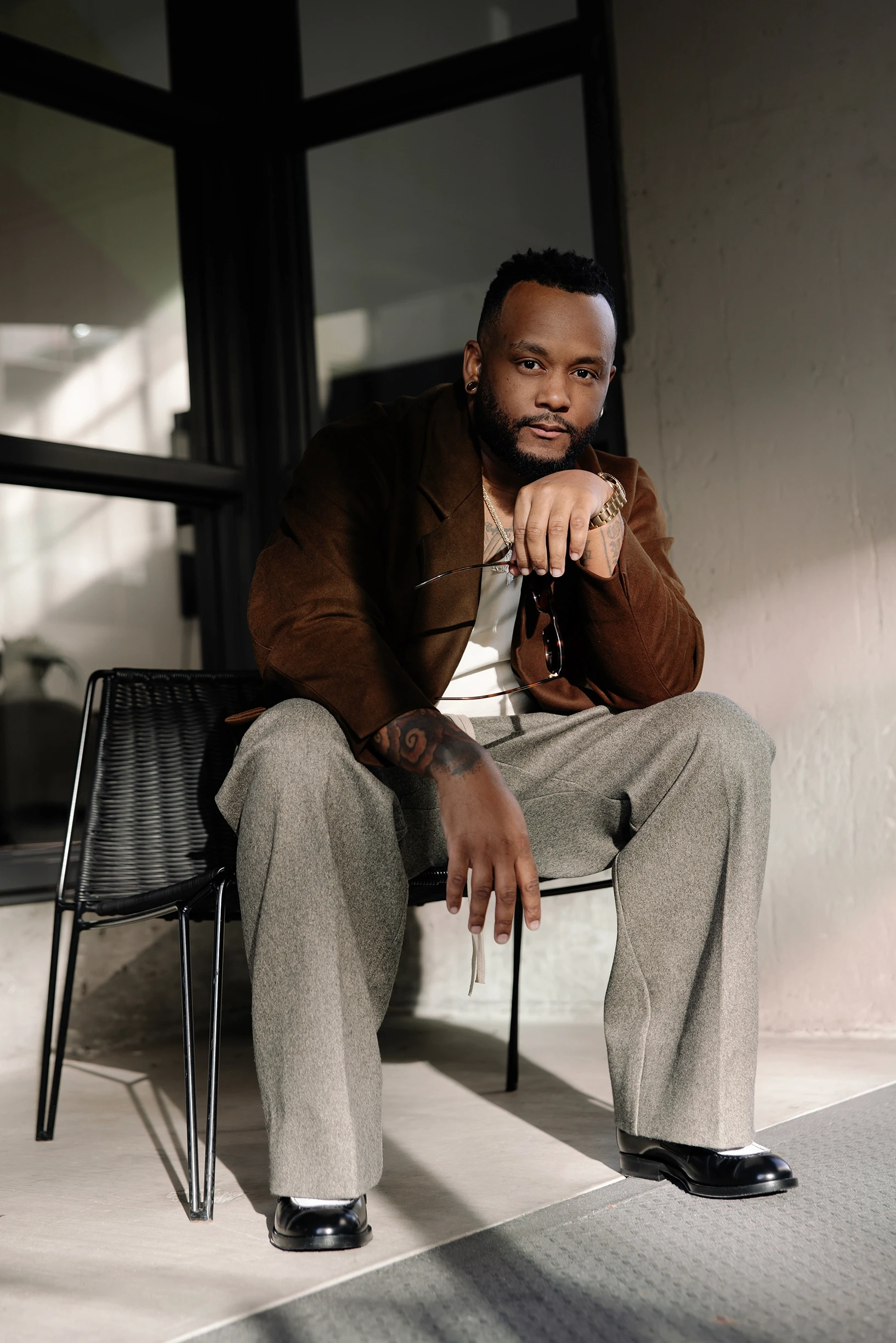 Rio Henderson headshot citting in a chair stylishly holding a pair of sunglasses. He is wearing a brown suede blazer, a white t shirt, a gold watch, heather gray pants, and black & white loafers