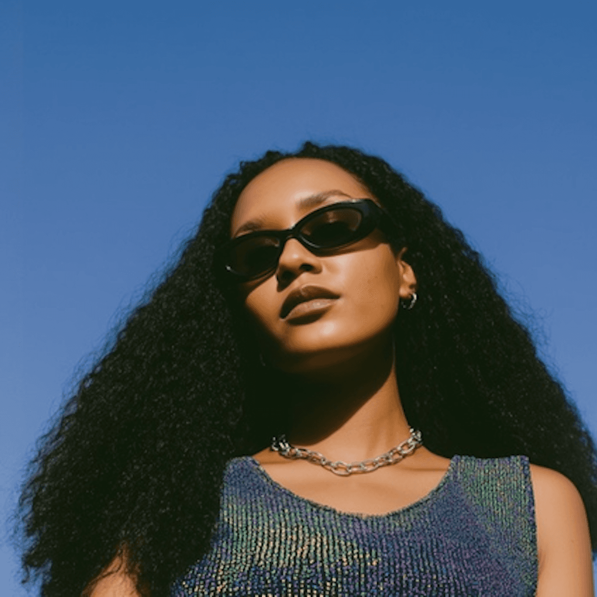 Confident woman with long curly hair wearing sunglasses and a shimmering top, photographed from a low angle against a deep blue sky.