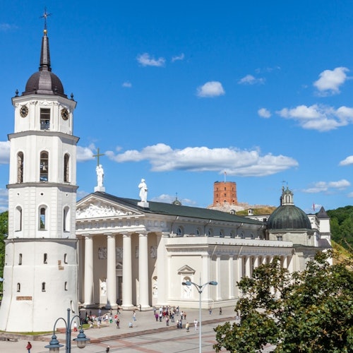 White cathedral with a tall bell tower and columns in front, people walking around, blue sky with scattered clouds above.