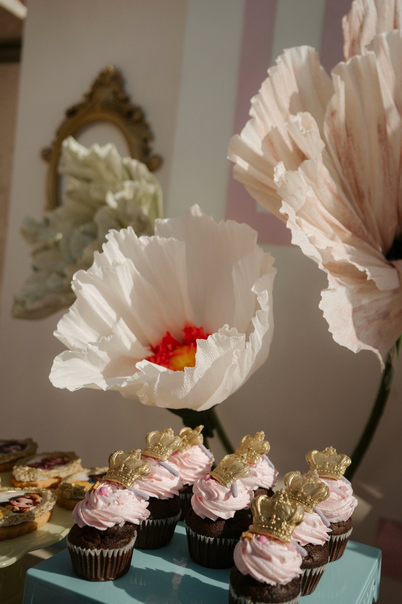 Pink-frosted cupcakes topped with gold crowns beneath oversized paper flowers at a princess-themed children’s party