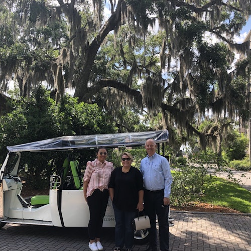 三个人站在一辆白色嘟嘟车前,车上有绿色座椅,车下是挂满西班牙苔藓的大树枝。